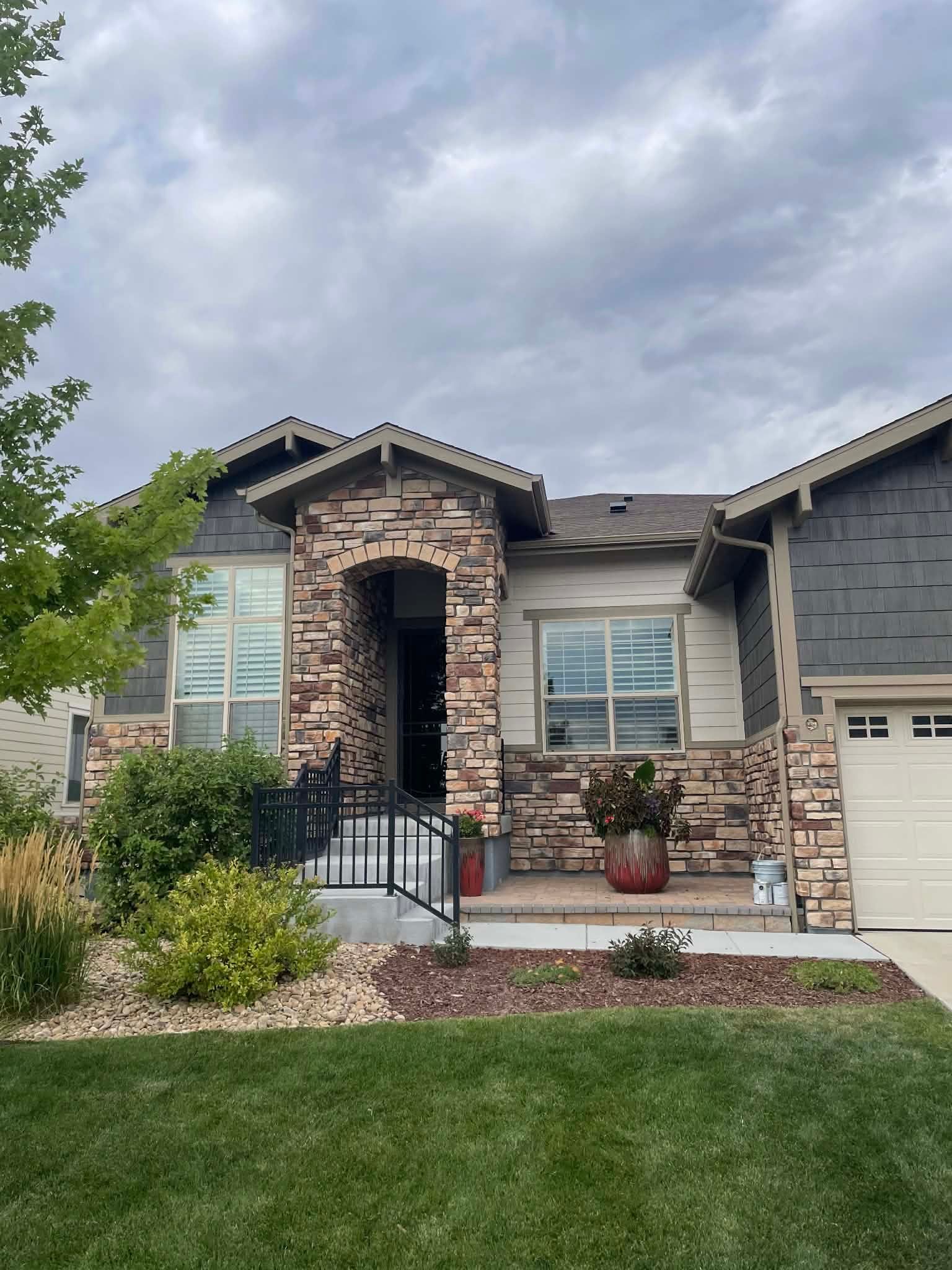 A residential house exterior with tan stone facade, gray siding, a covered front porch, and a white garage door.