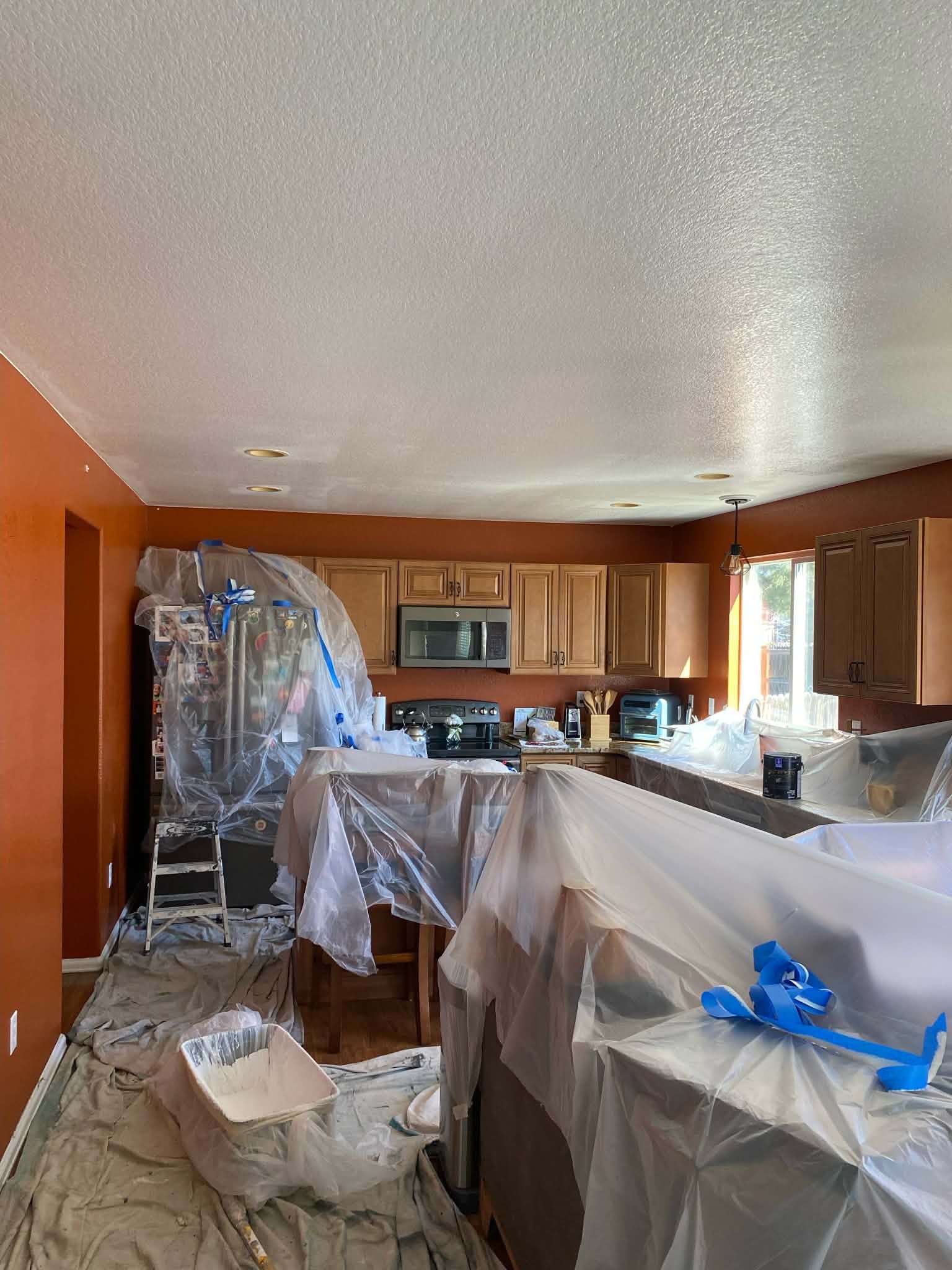 A kitchen undergoing painting preparation with surfaces and appliances covered in plastic drop cloths.