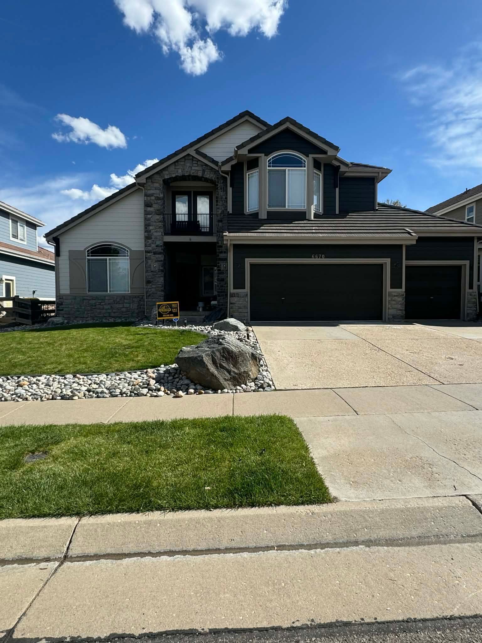 A two-story suburban house with dark gray siding, stone accents, a multi-car garage, and a lawn under a blue sky.