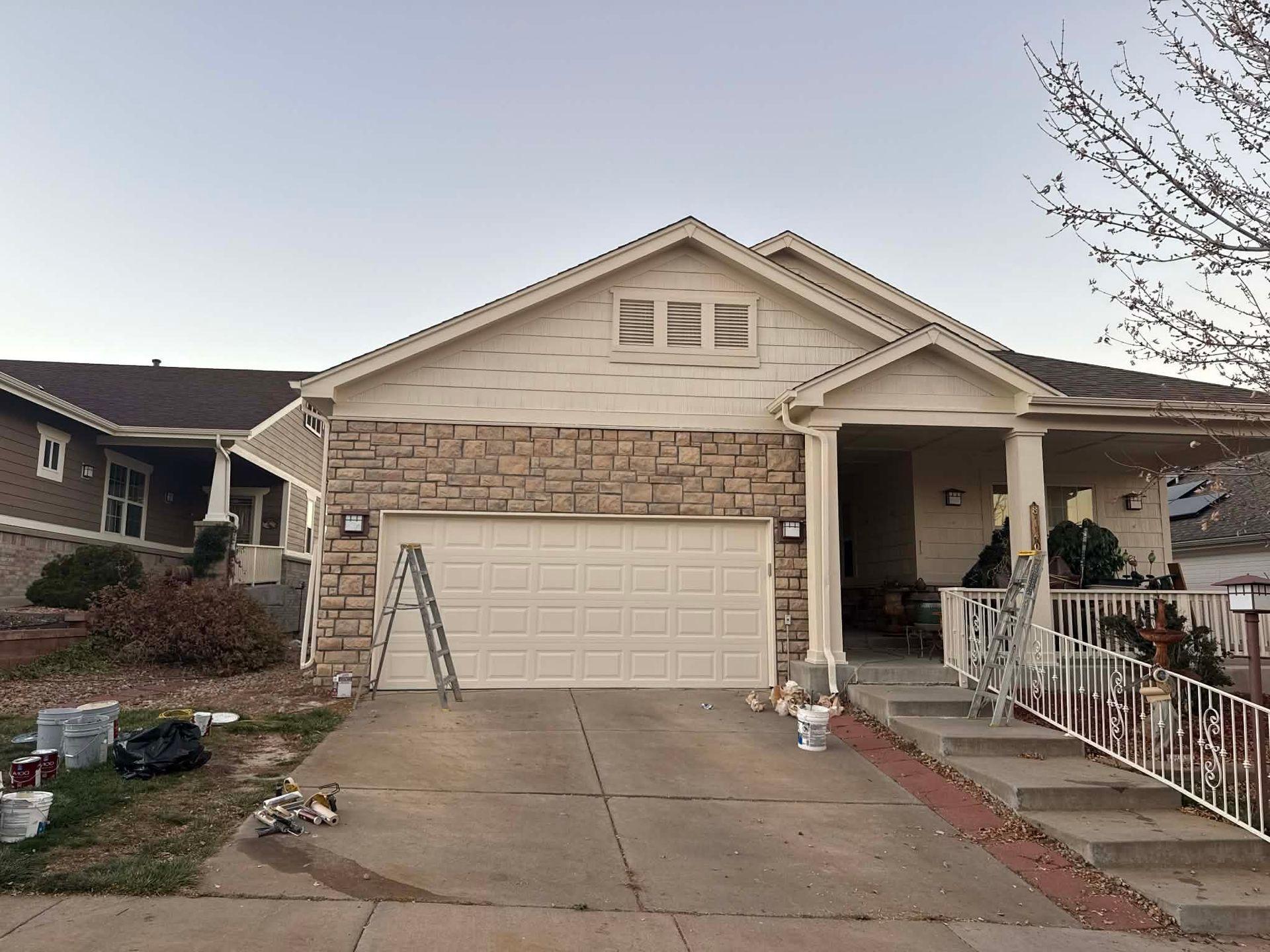 A one-story beige house with a stone facade, a two-car garage, a porch with white railings, and a ladder in the driveway.