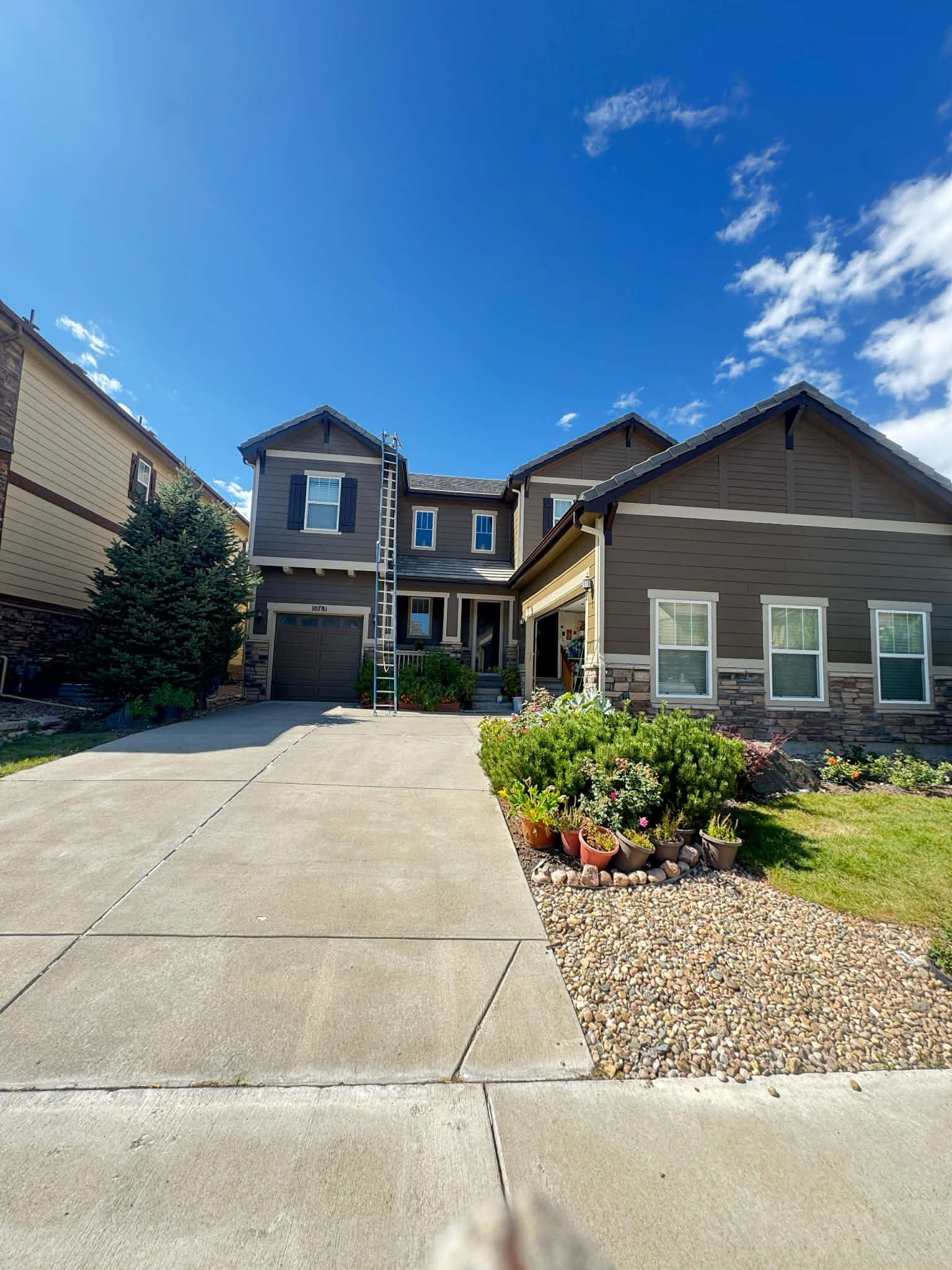 A two-story brown house with a concrete driveway, a small garden, and a clear blue sky.