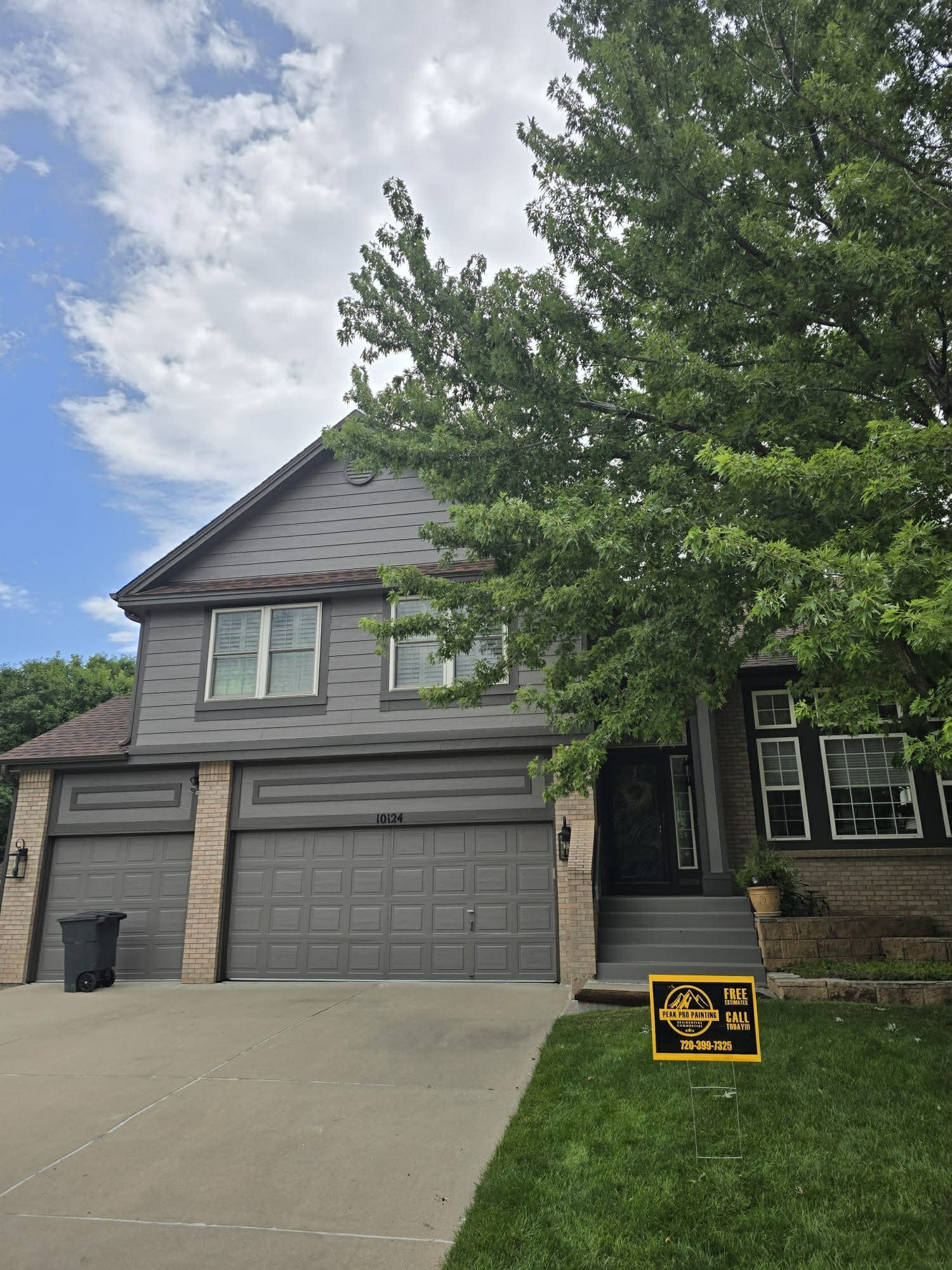 Two-story gray house with a garage, concrete driveway, and a large tree in the front yard under a partly cloudy sky.