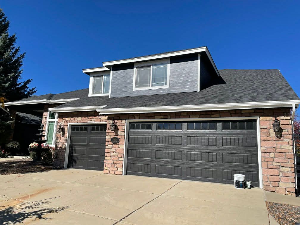 A house with a stone facade, a gray shingle roof, and two dark garage doors, situated under a clear blue sky.