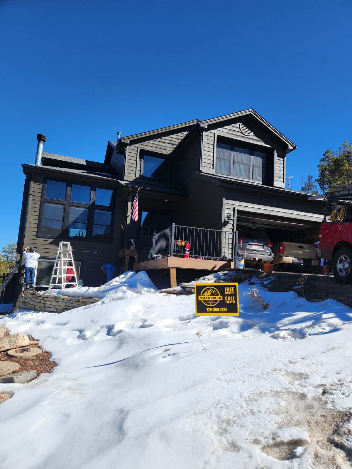A dark-colored, multi-story house sits on a snowy lot with a ladder, vehicles, and a yellow construction sign in front.