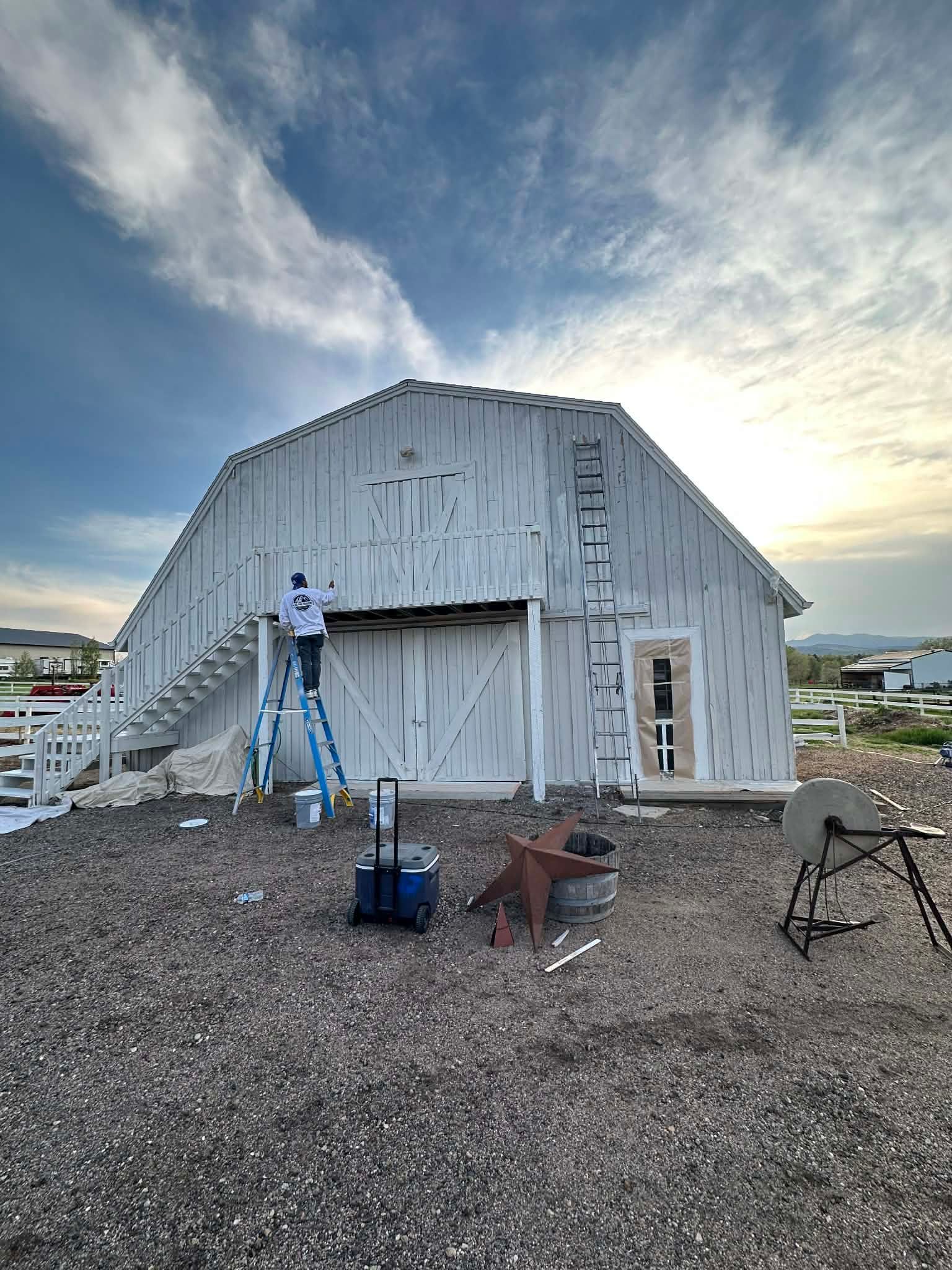A person on a ladder paints a white barn under a cloudy sky. A cooler and a metal star sit on the rocky ground nearby.