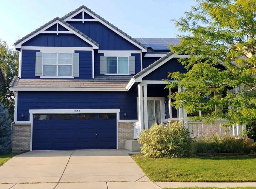 A two-story blue house with a two-car garage, gray shutters, and a front porch, set on a suburban lawn.