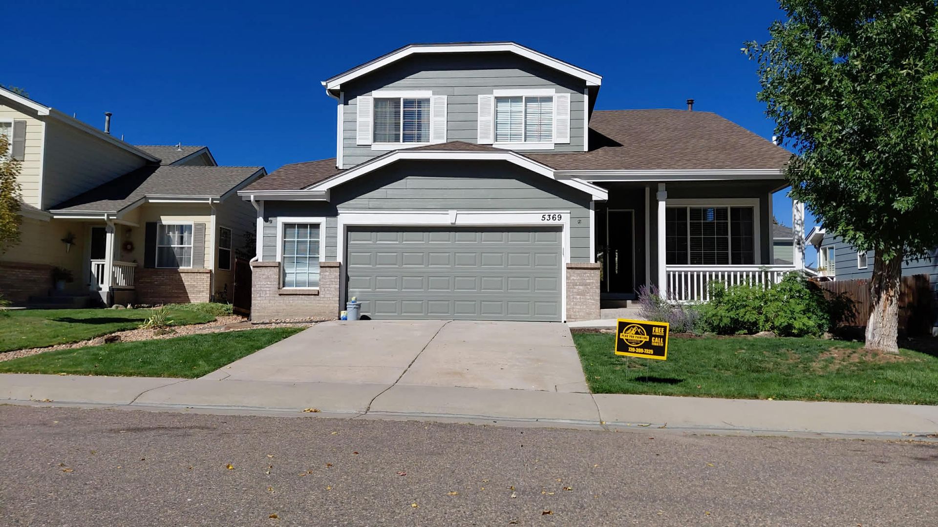A gray, two-story suburban house with a two-car garage, front porch, and a yellow real estate sign on the front lawn.
