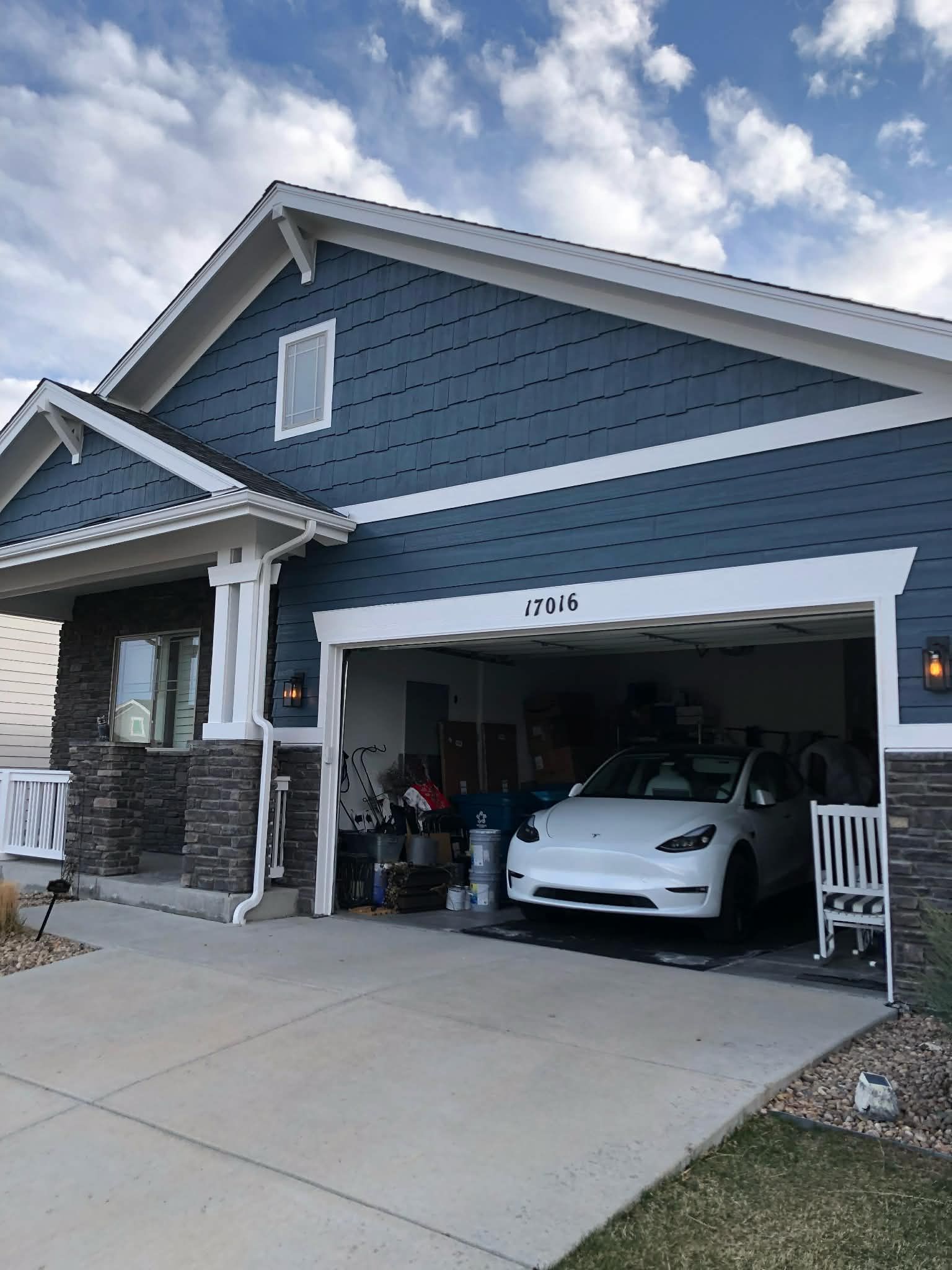 A blue house with a stone base and an open garage containing a white Tesla car on a sunny day.