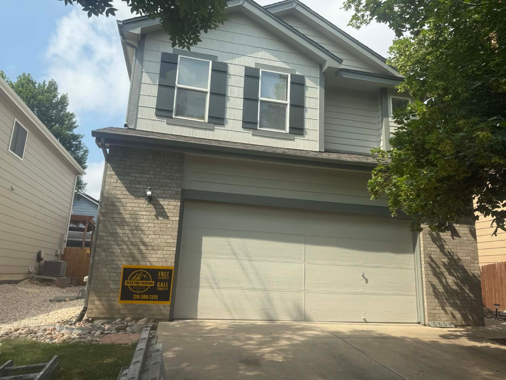 A two-story gray house with a stone facade, a two-car garage, and a yellow sign on the front corner.