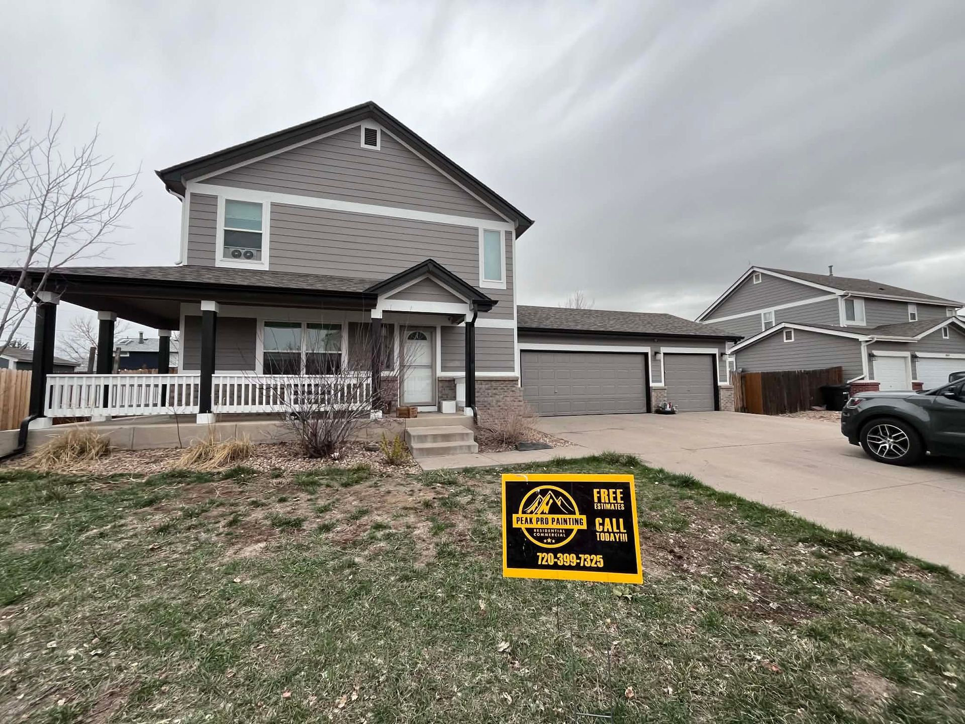 A two-story grey suburban house with a wrap-around porch and a three-car garage, featuring a yellow real estate yard sign.