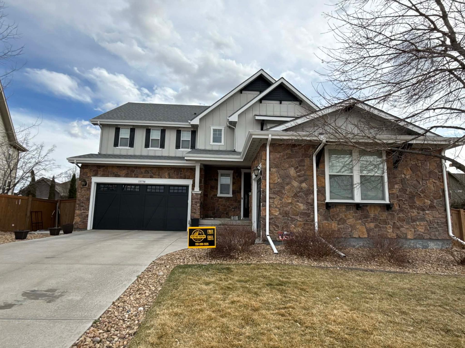 Two-story house with a stone facade, beige siding, a black two-car garage, and a sign on the front lawn.