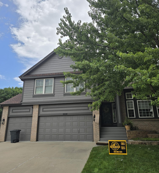 A two-story grey house with a two-car garage, a tree in the front yard, and a yellow real estate sign on the lawn.