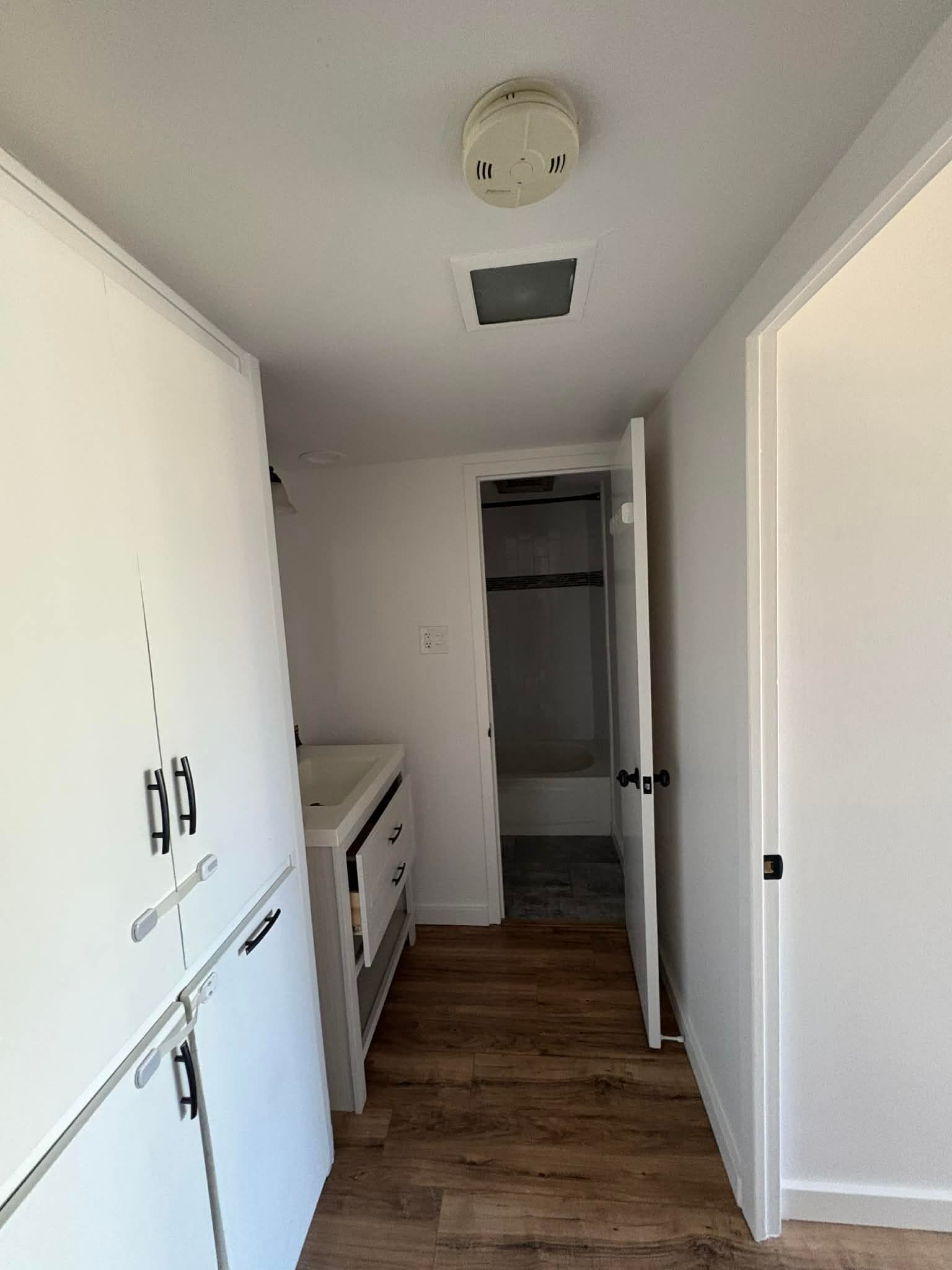 A hallway with white walls and wood-look flooring leading to a bathroom doorway with a vanity and shower visible.