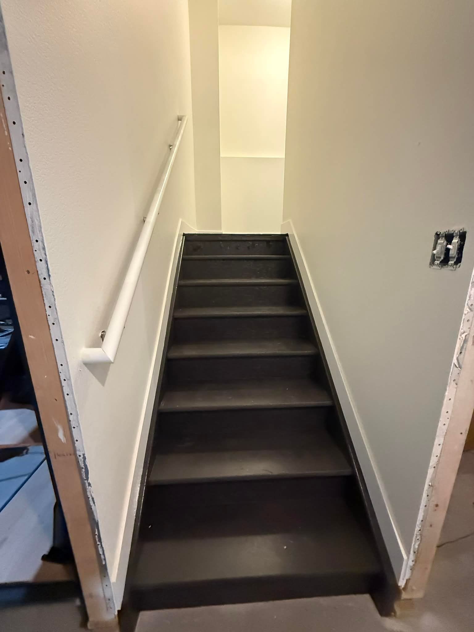A straight, indoor staircase with dark treads leading upward between two light-colored walls, featuring a white handrail.