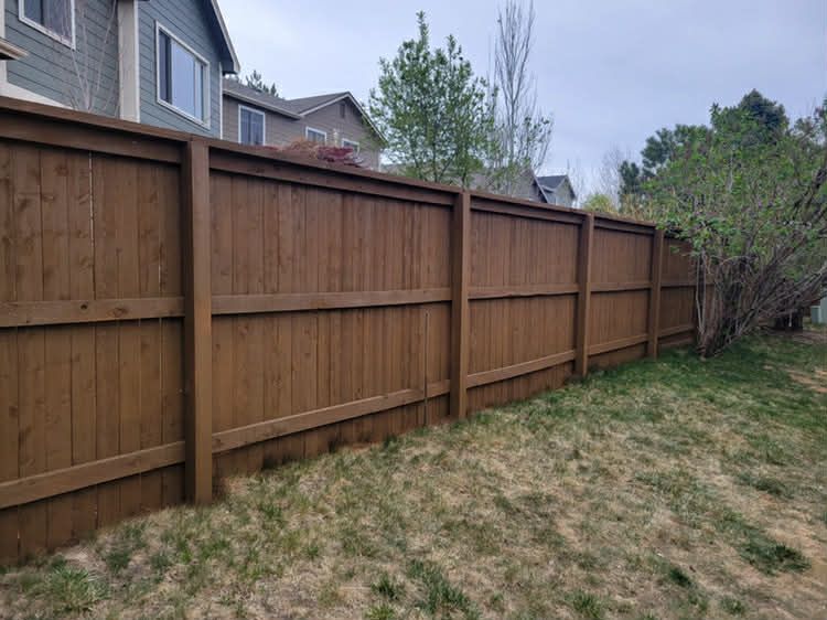 A long, brown wooden privacy fence running alongside a grassy yard with houses in the background.
