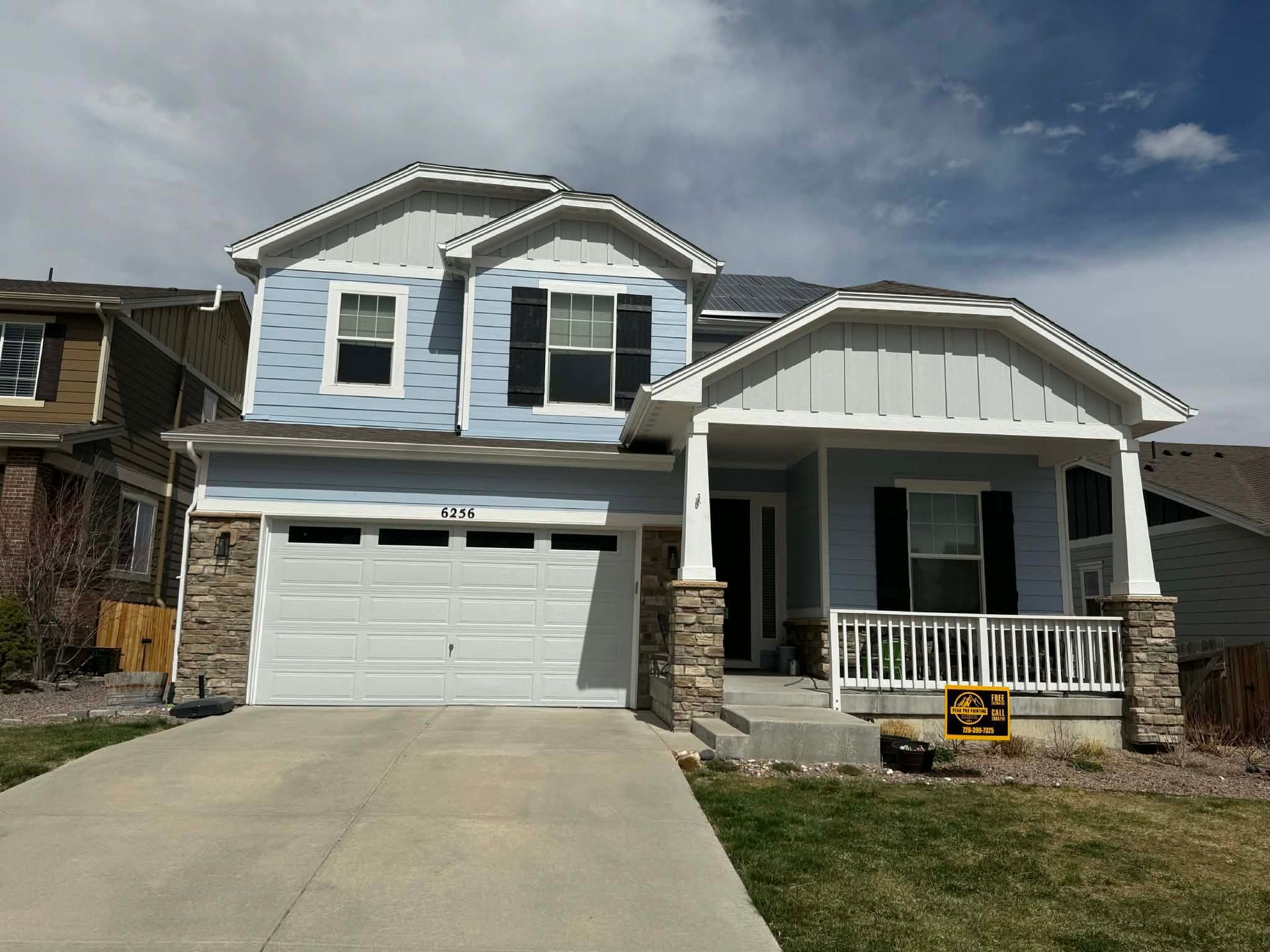 A two-story light blue house with a two-car garage, stone accents, and a front porch under a partly cloudy sky.