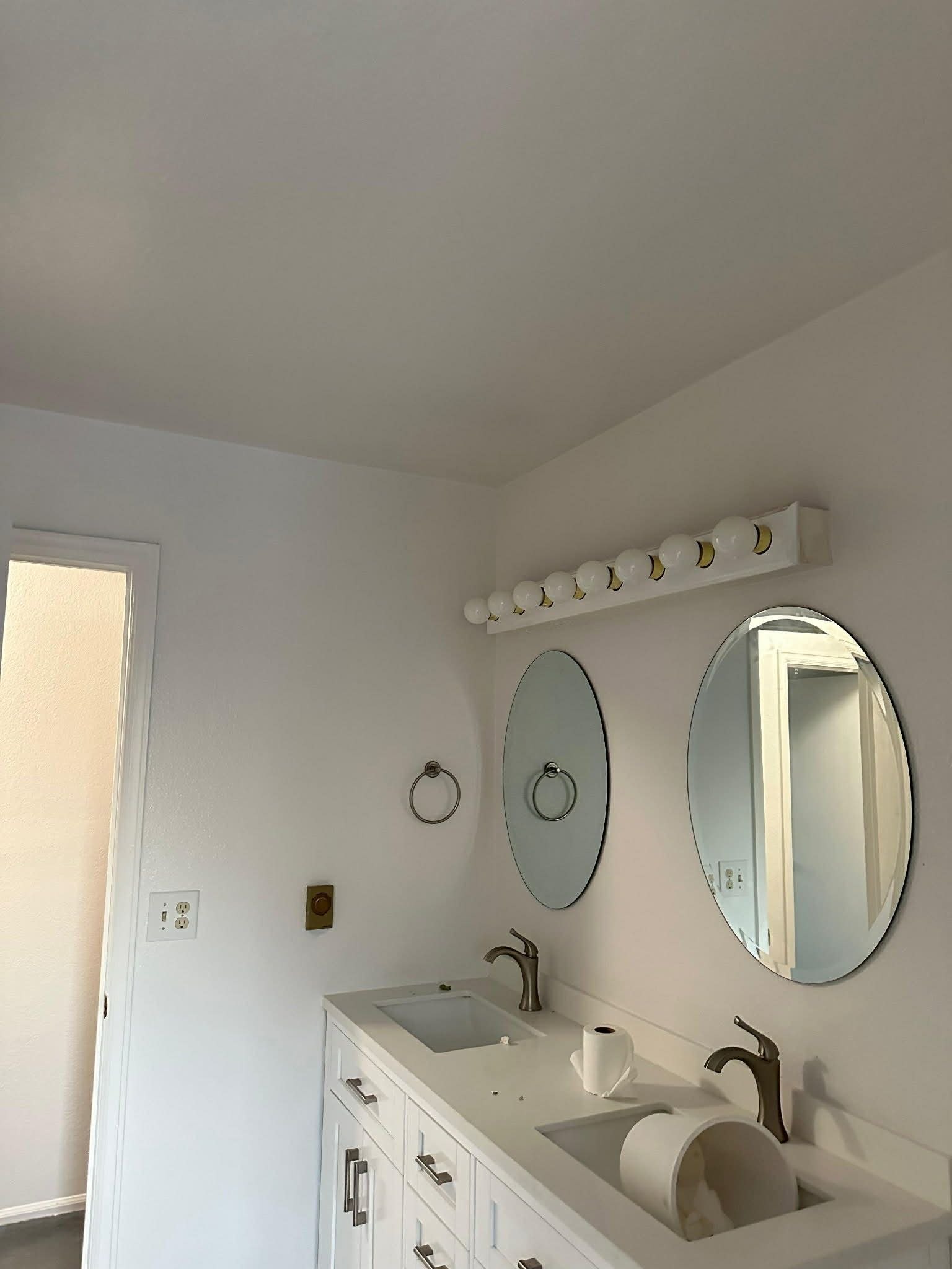 A bright, white bathroom vanity with double oval mirrors, gold faucets, and a Hollywood-style light fixture above.