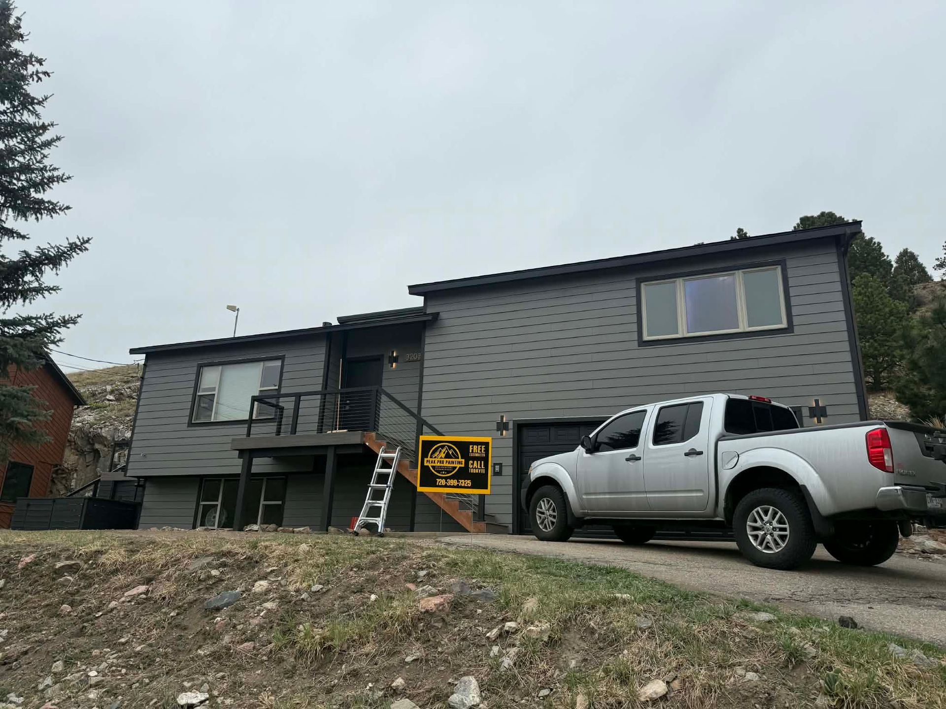 A grey two-story house with a ladder leaning against the porch and a silver pickup truck parked in the driveway.