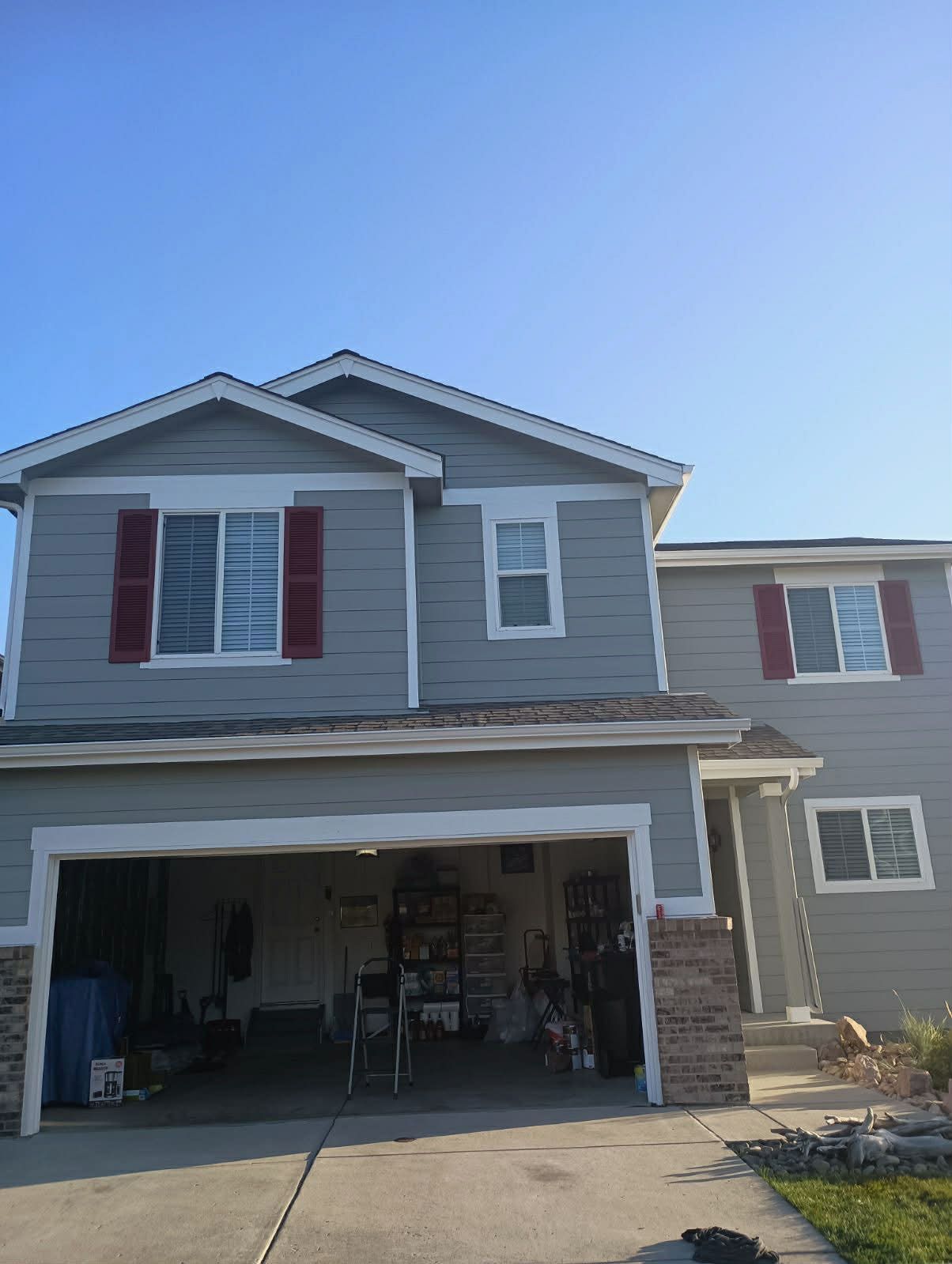 A two-story grey suburban house with red window shutters and an open two-car garage under a clear blue sky.