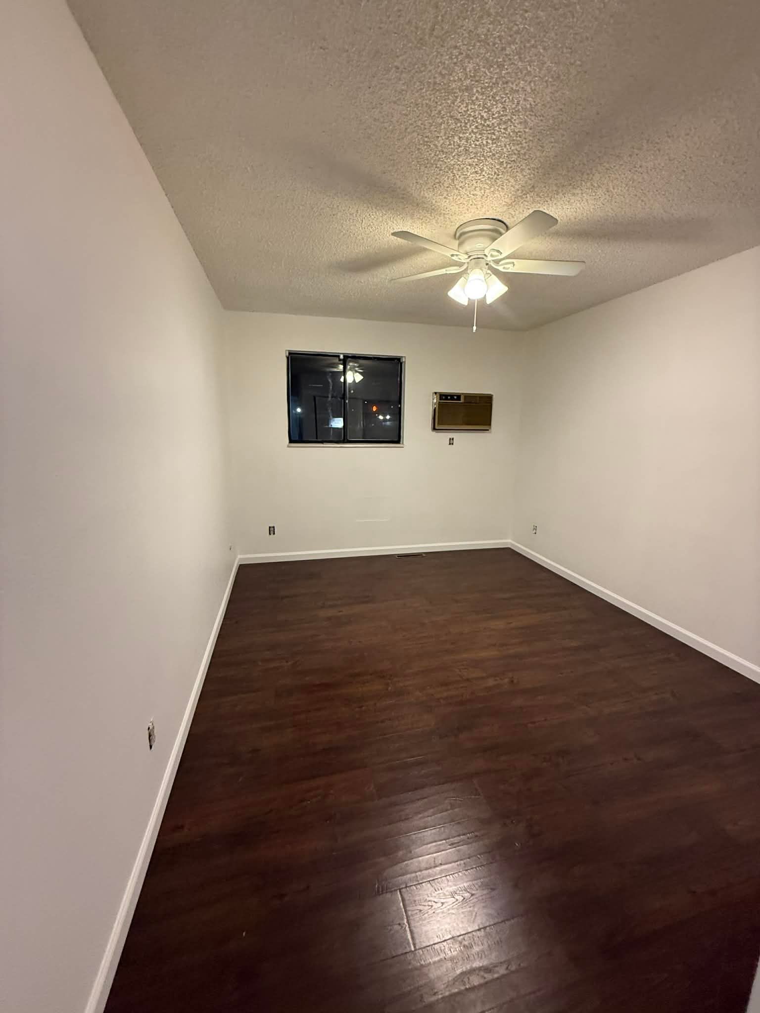 An empty room with dark wood floors, white walls, a ceiling fan, a window, and a wall-mounted air conditioner.