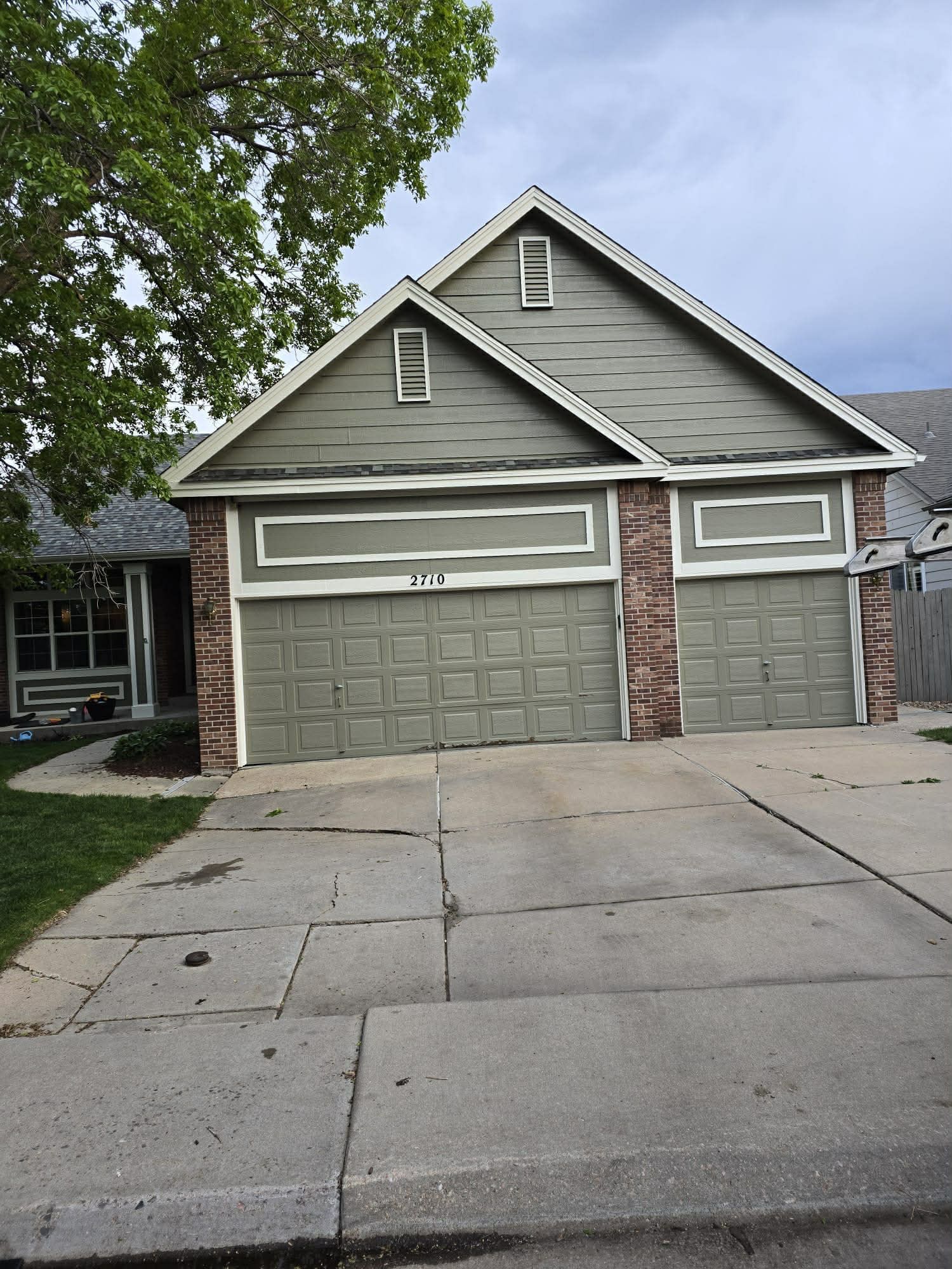A green-sided house with a large garage, brick accents, and a concrete driveway under an overcast sky.