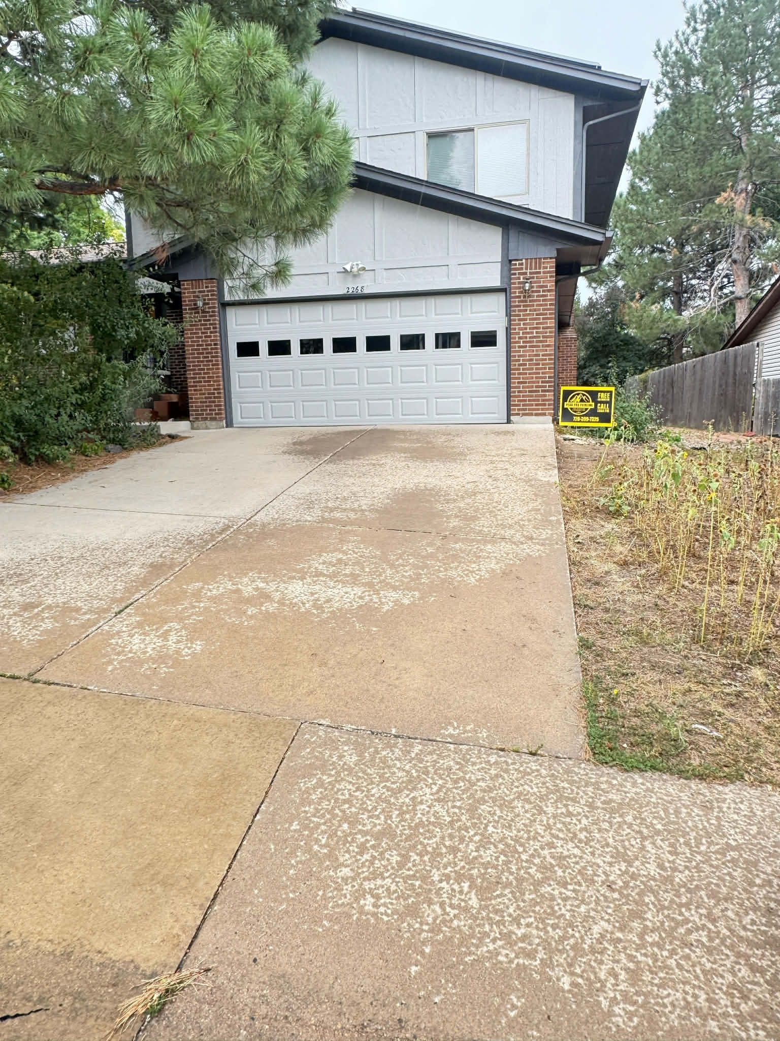 Two-story house with gray vertical siding, brick accents, and a white garage door, seen from a paved driveway.