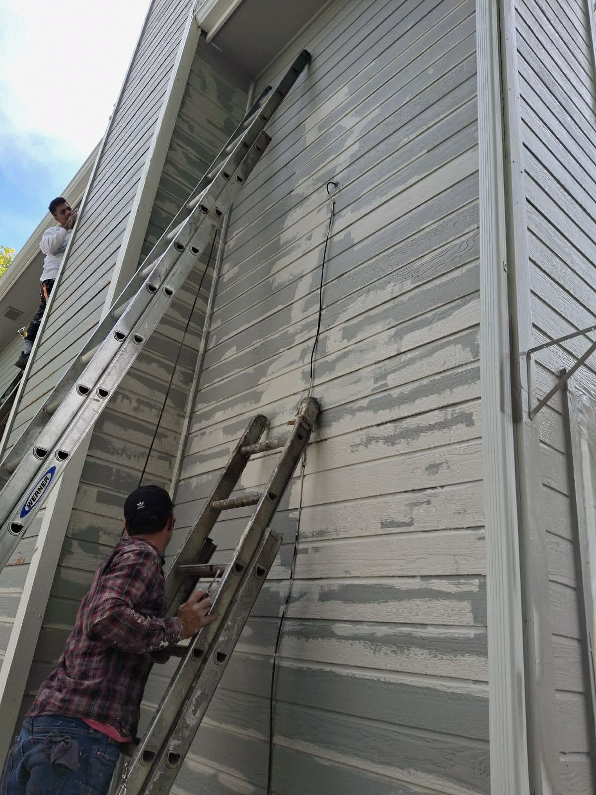 Two workers on extension ladders are repairing or painting the light gray wooden siding of a multi-story building.