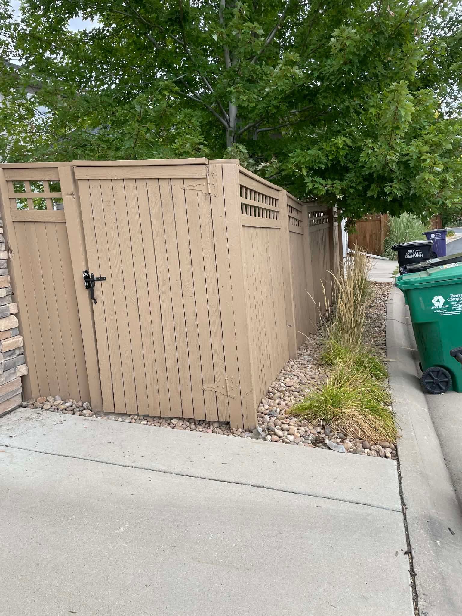 A tan wooden privacy fence enclosure for trash cans stands on a gravel patch next to a concrete sidewalk.