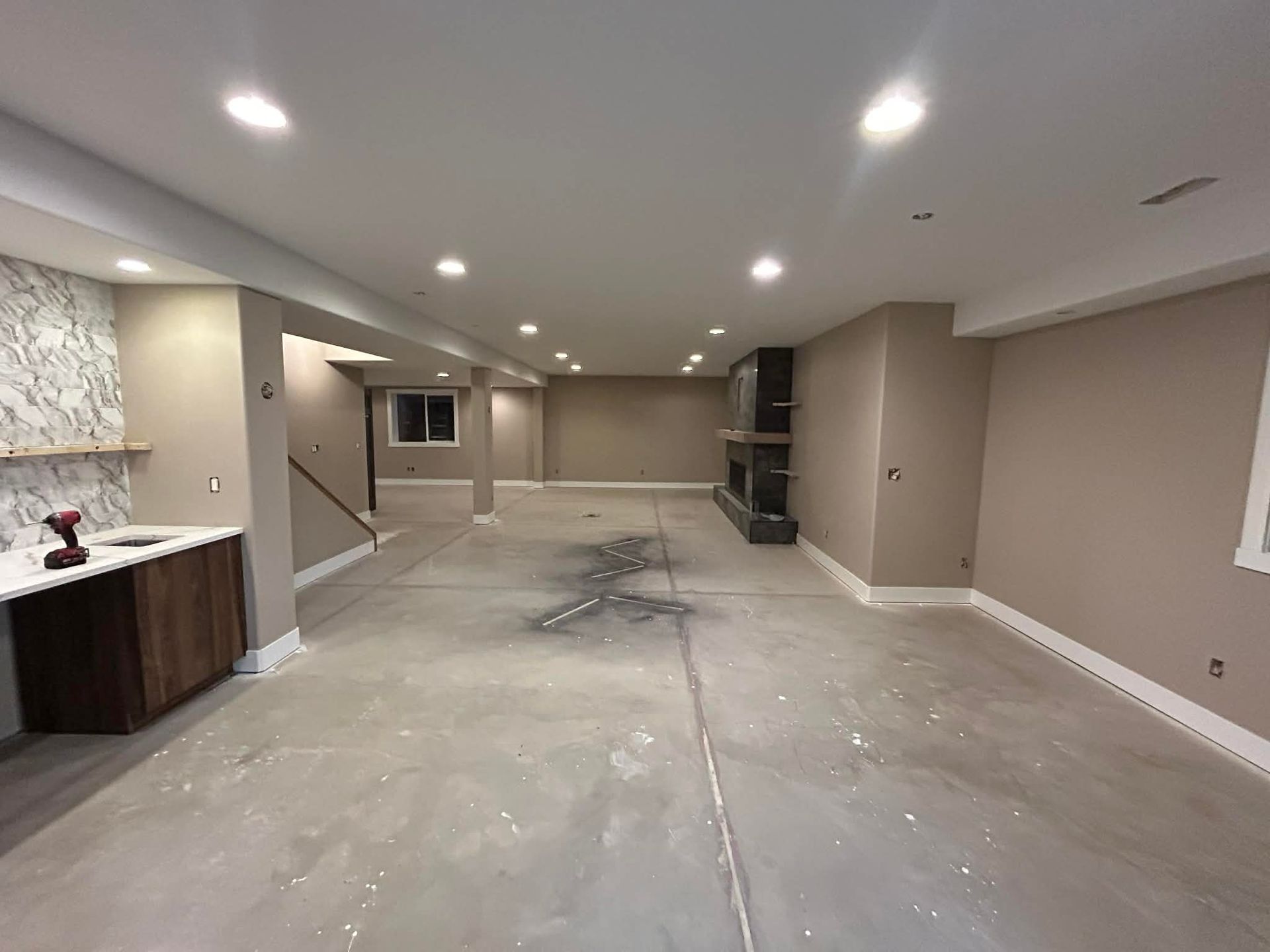 A wide, unfinished basement with beige walls, recessed lighting, a marble-topped kitchenette, and a stone fireplace.