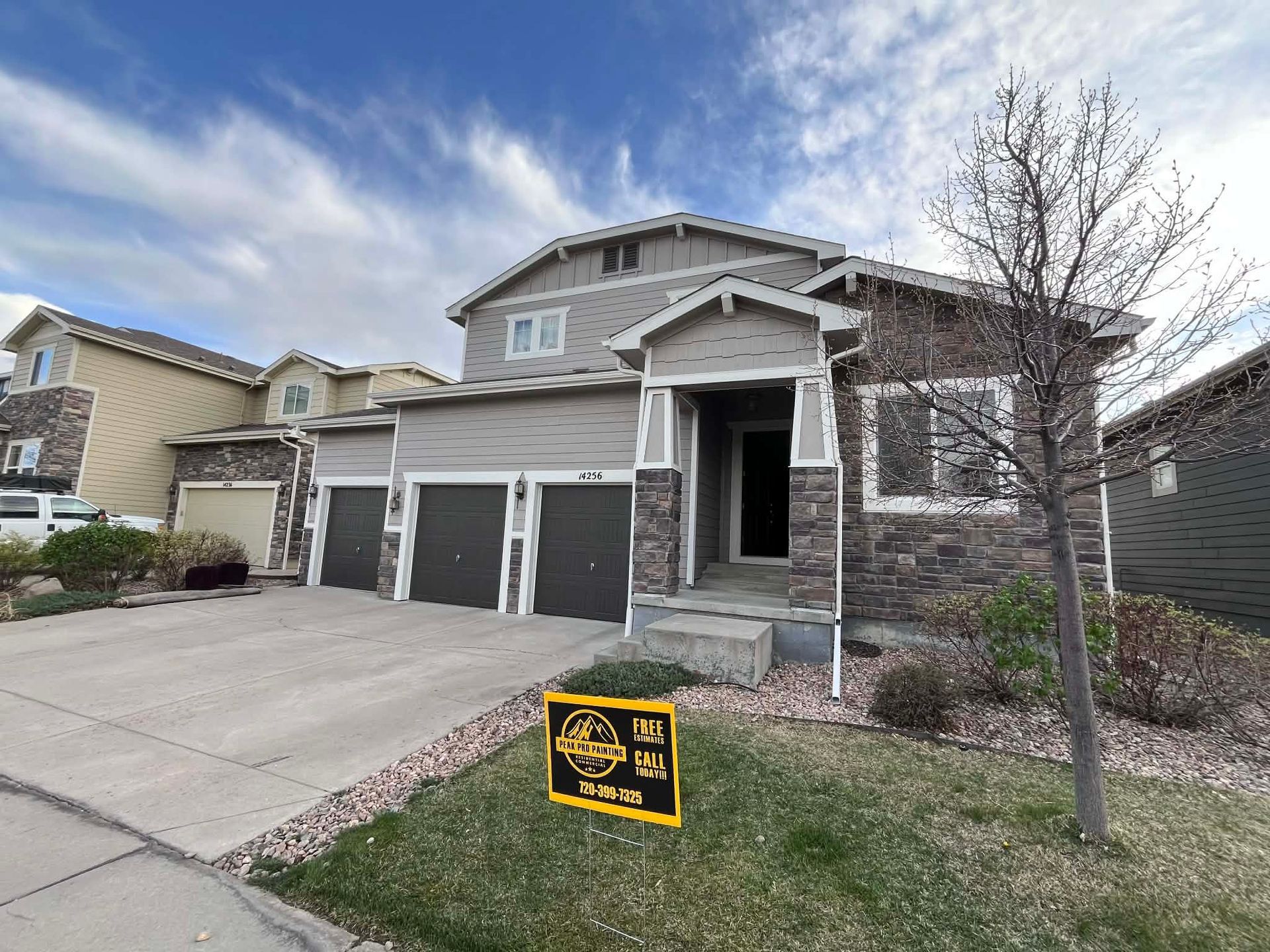 A two-story suburban house with stone and grey siding, a three-car garage, and a yard sign, under a cloudy blue sky.
