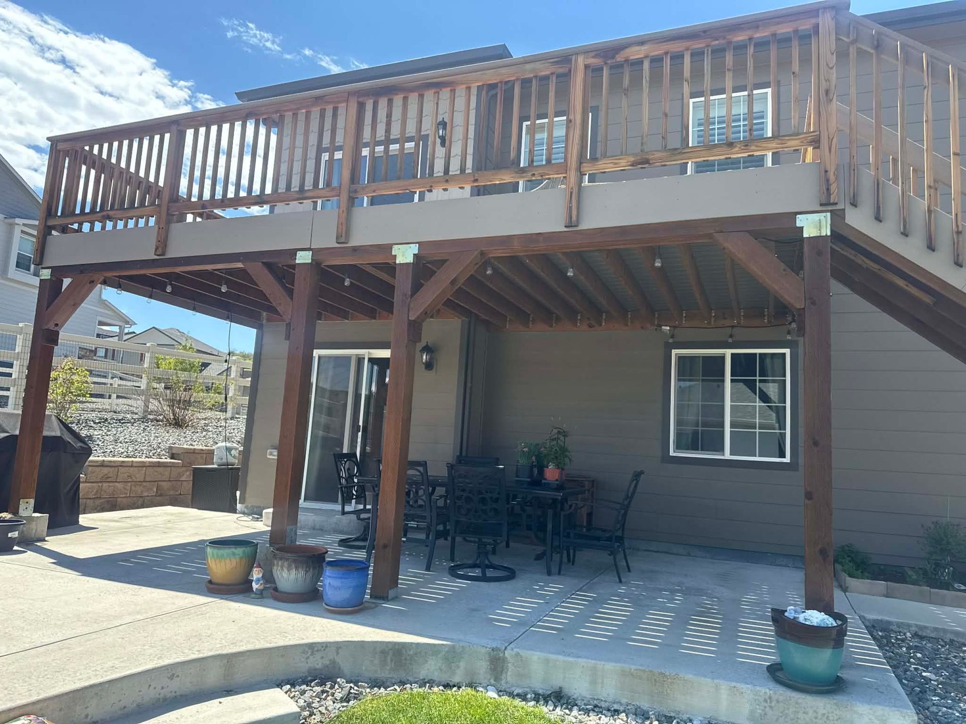 A taupe house with a large wooden deck above a concrete patio, furnished with chairs and potted plants.