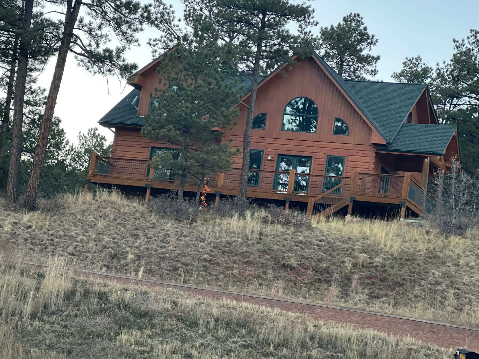 A large, multi-story wooden cabin with a dark shingled roof sits on a grassy hillside surrounded by tall pine trees.