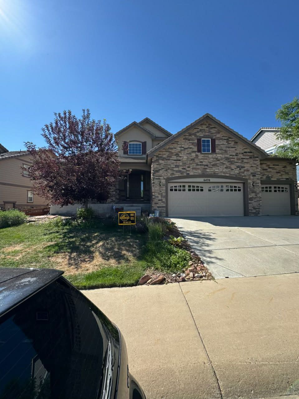 A tan stone house with a three-car garage and a small tree in the front yard under a clear blue sky.