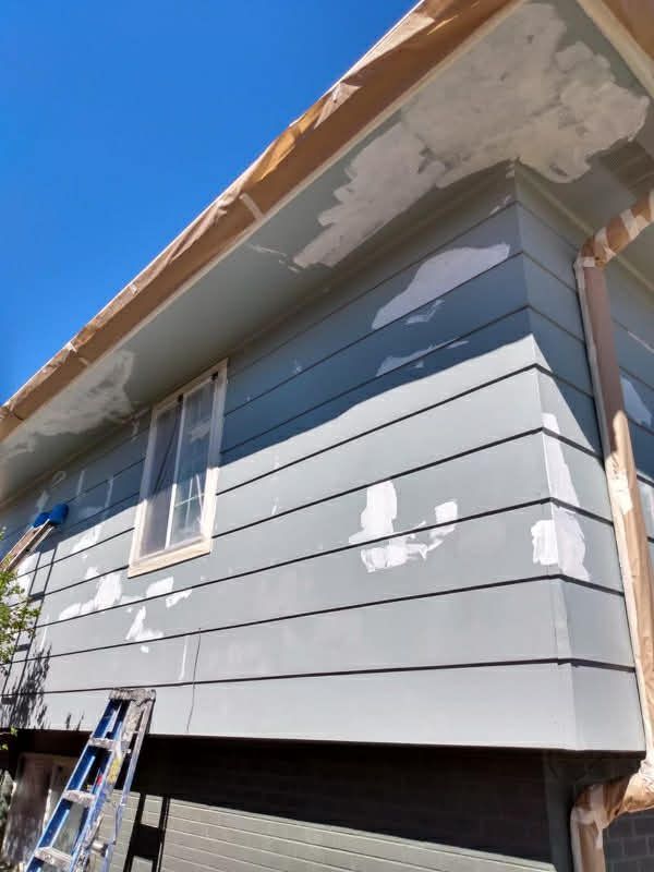 Exterior wall with blue siding featuring patches of exposed primer under the eaves and near a window, with a ladder below.