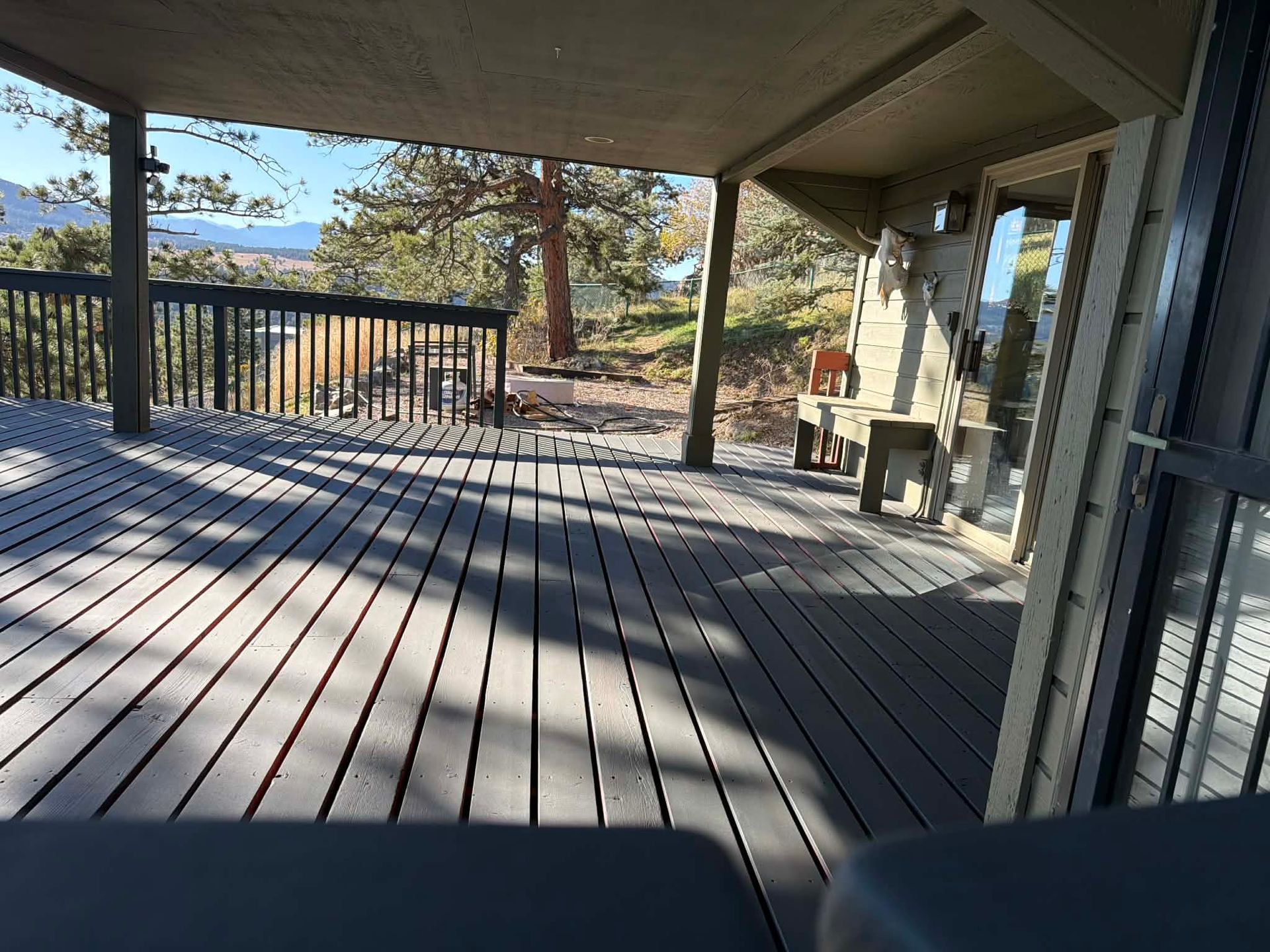 A spacious outdoor wooden deck under a covered patio, featuring a railing, a small bench, and a view of trees and mountains.
