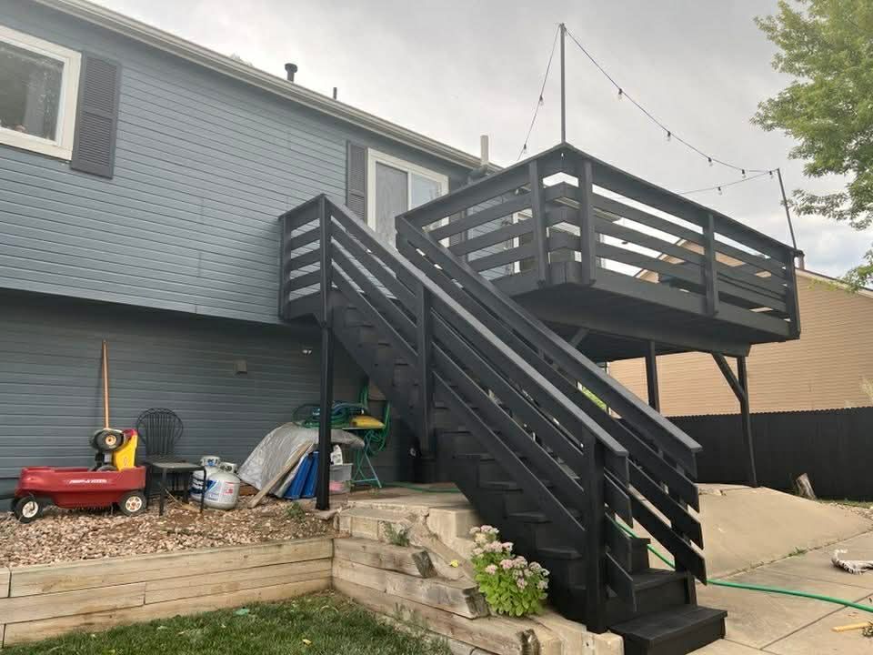 A black wooden deck with stairs attached to the back of a grey house, positioned above a patio and a retaining wall.
