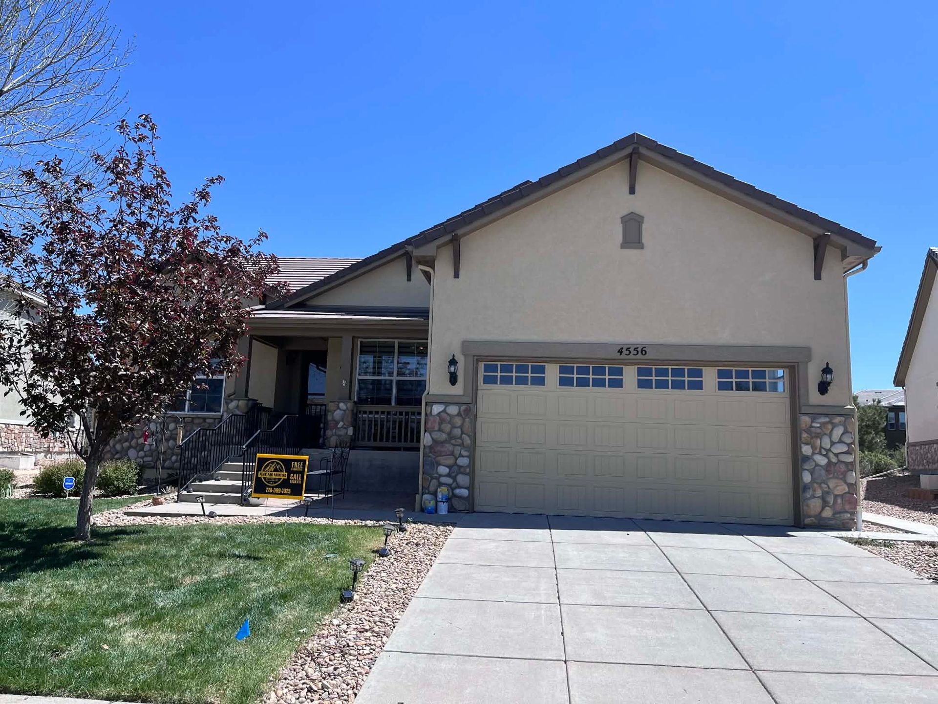 A one-story suburban home with tan stucco, stone accents, a two-car garage, and a green lawn under a clear blue sky.