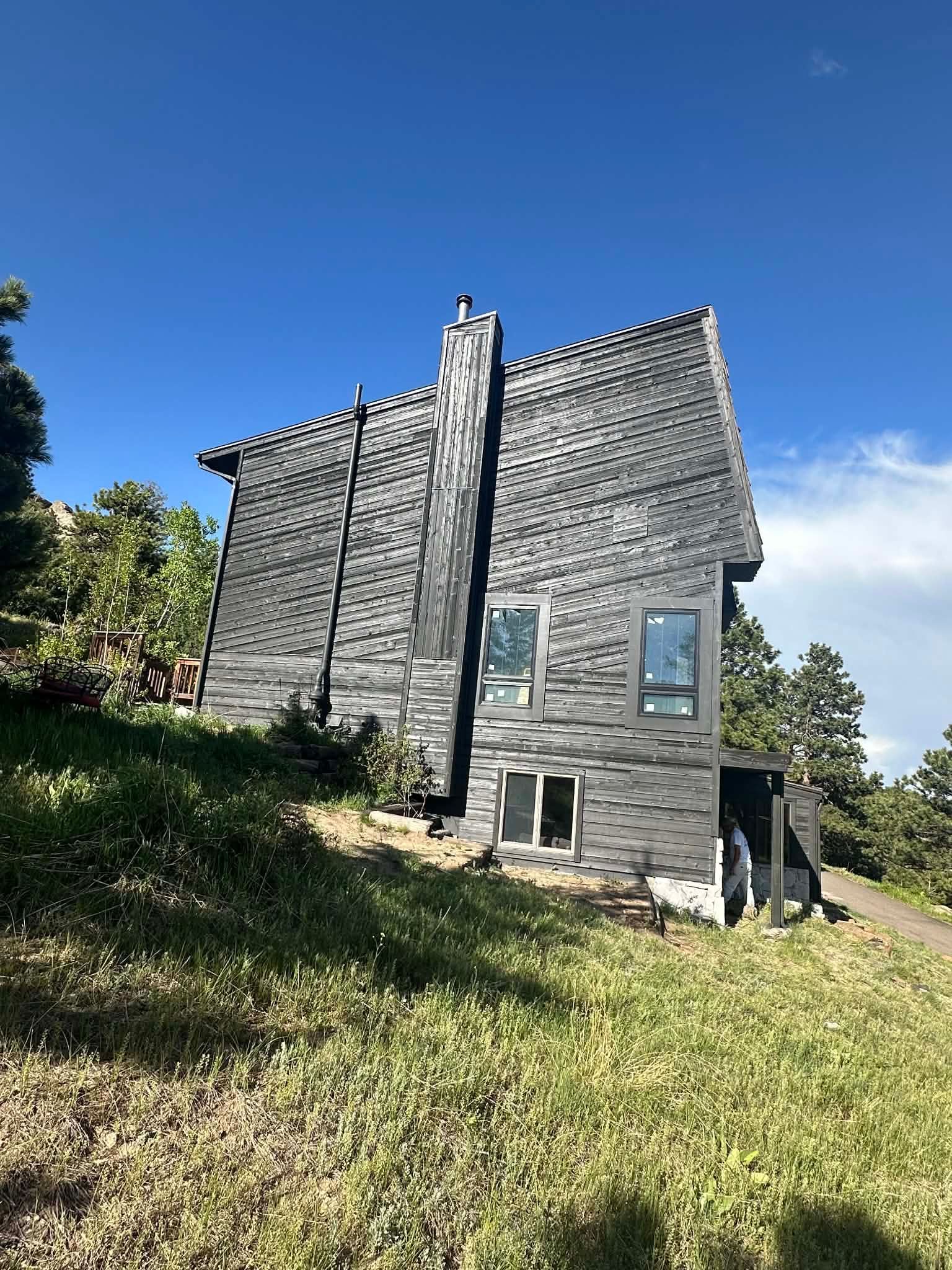 A modern, dark-sided house with a central chimney stands on a grassy, sunlit hillside under a bright blue sky.