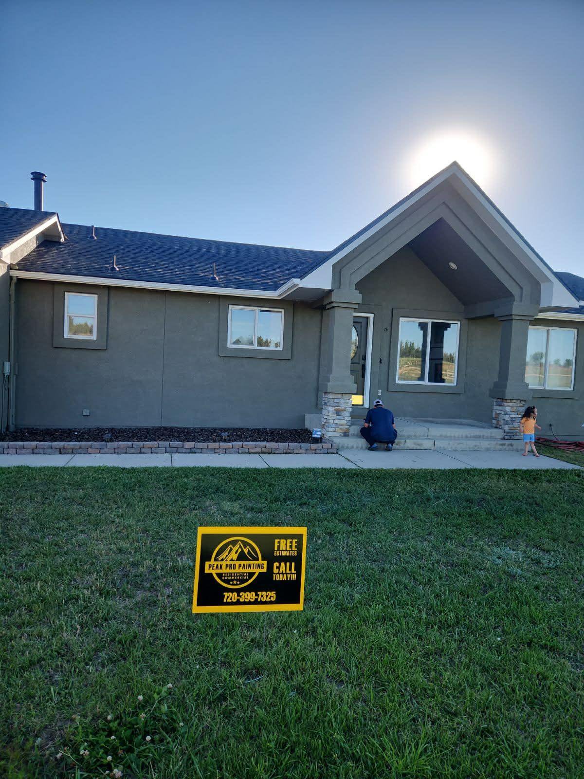 A gray house with a peaked roof and a yard sign in the grass. A person is sitting on the front steps in the bright sun.