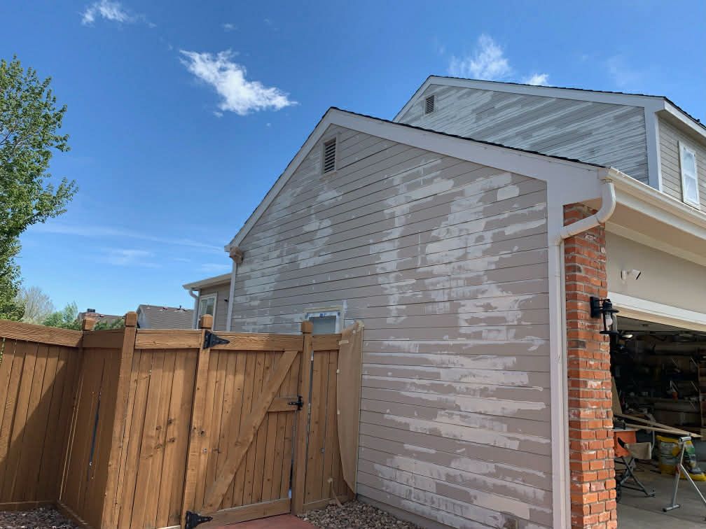 A two-story house exterior showing severe paint peeling on the light-colored siding, next to a wooden fence.