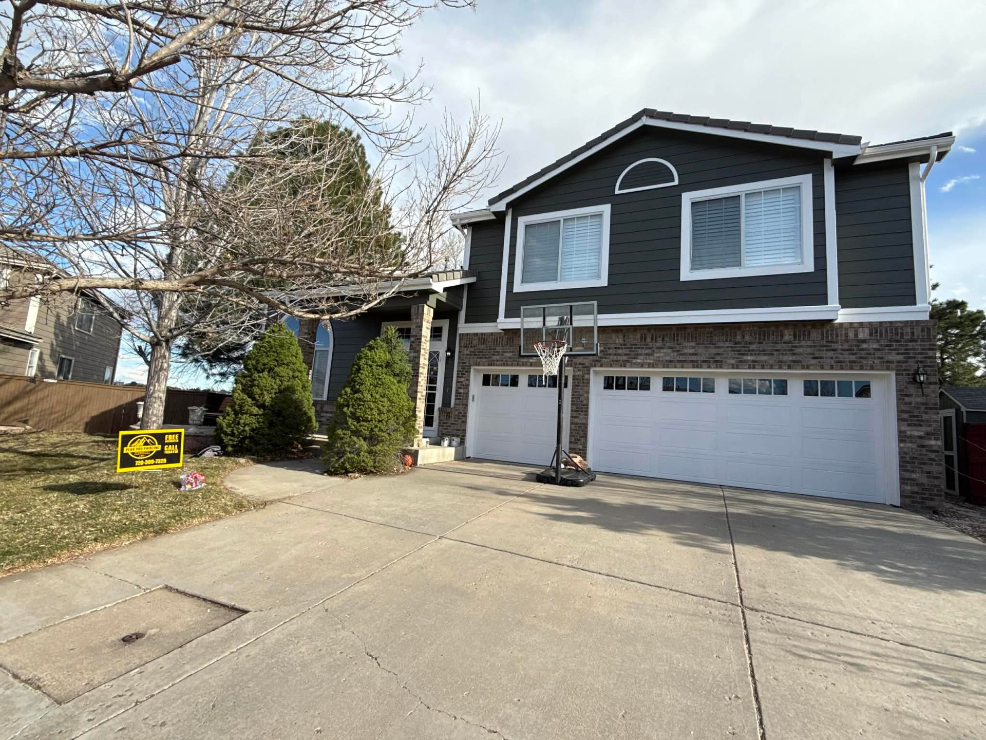 A two-story house with dark gray siding, stone accents, and a two-car garage, with a driveway and a bare tree out front.