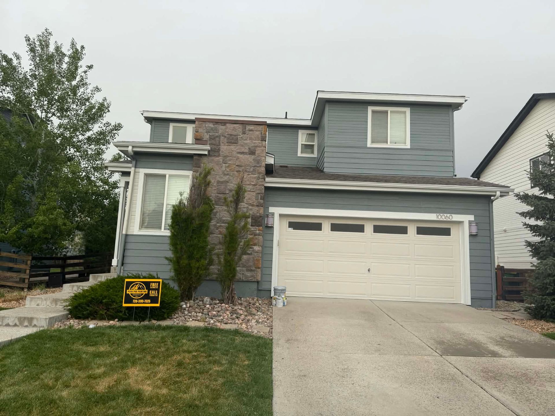 A two-story house with grey siding, a stone facade chimney, and a white two-car garage on a suburban street.