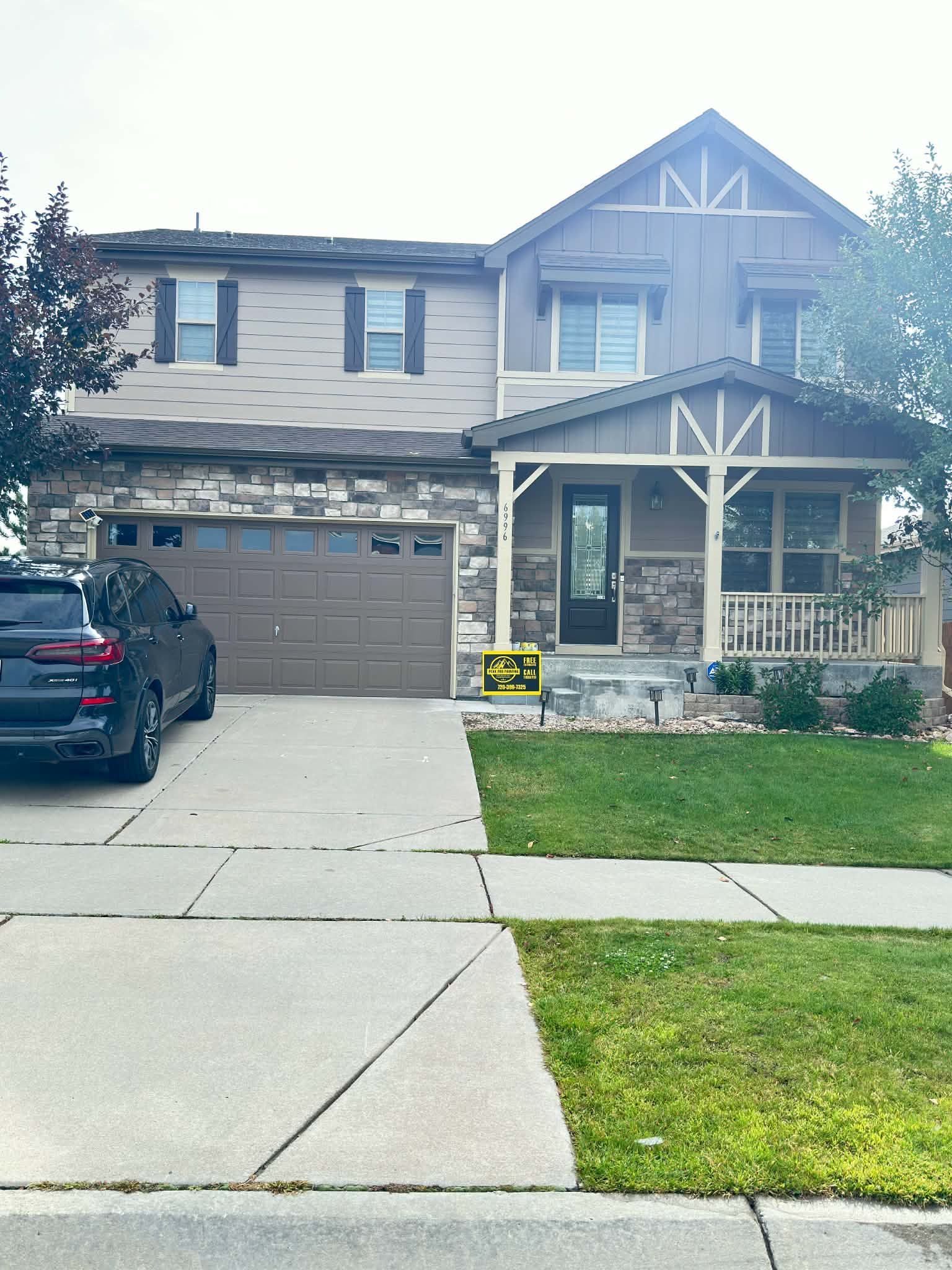 A two-story suburban house with stone accents, tan siding, a brown garage door, and a front porch, viewed from the street.