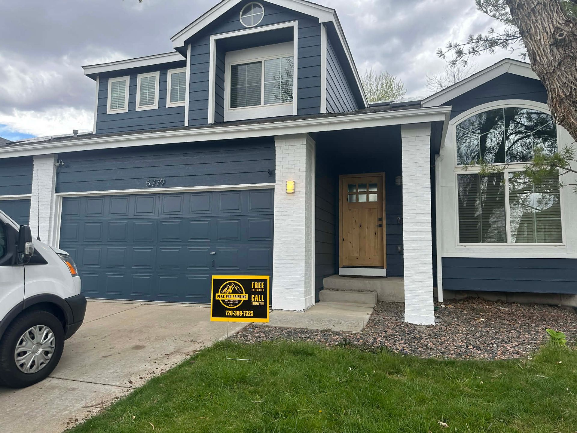 A two-story blue house with white trim and brick accents, featuring a wooden front door and a two-car garage.