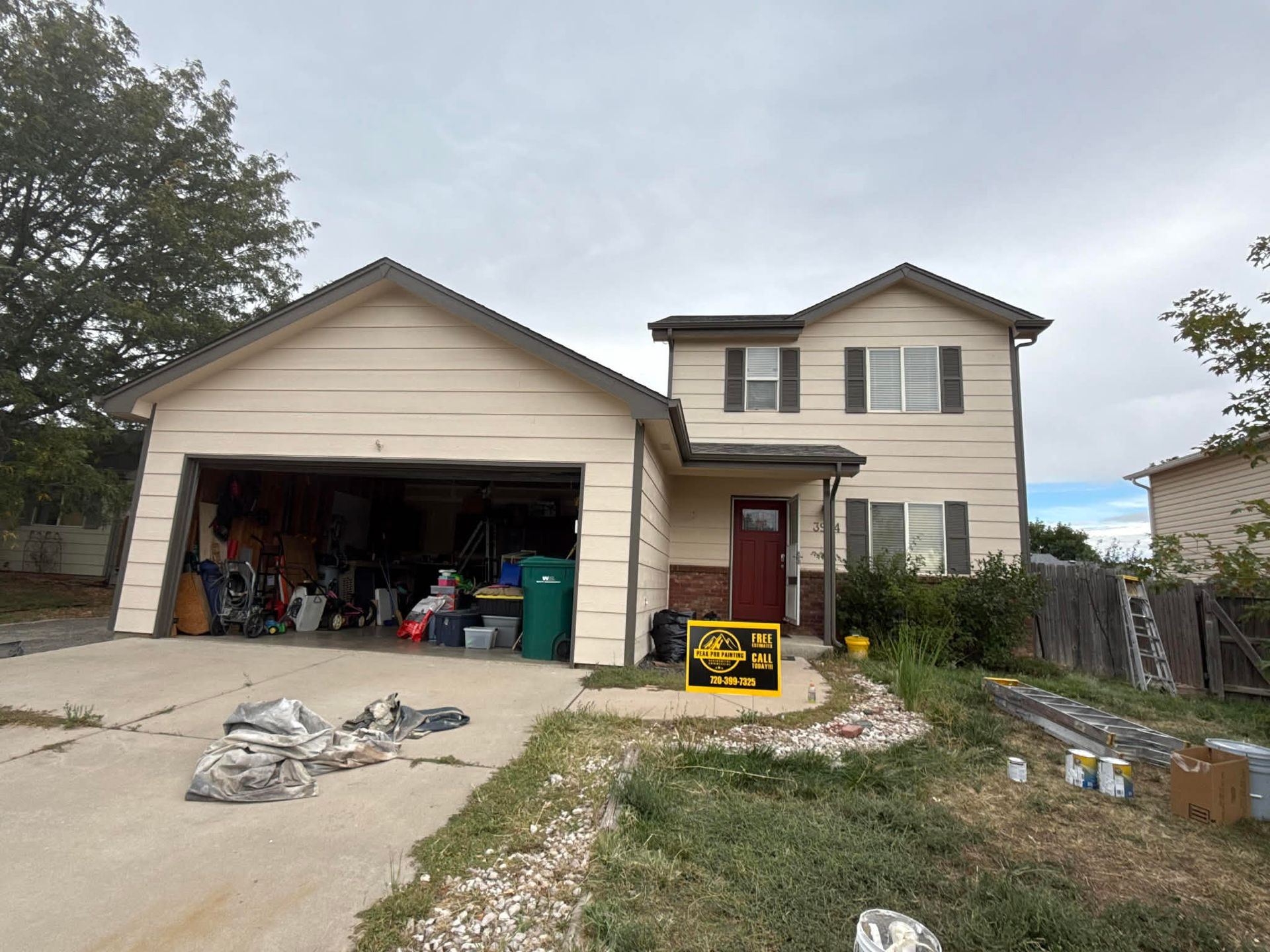 A two-story suburban house with beige siding, a garage, a red front door, and a yellow sign on the front lawn.