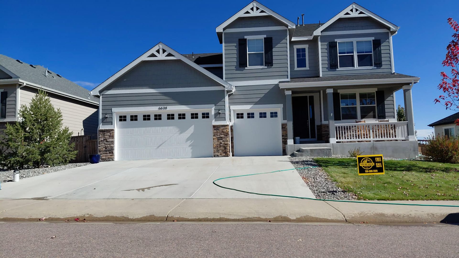 A two-story grey suburban house with white garage doors and a front porch, viewed from the street on a sunny day.