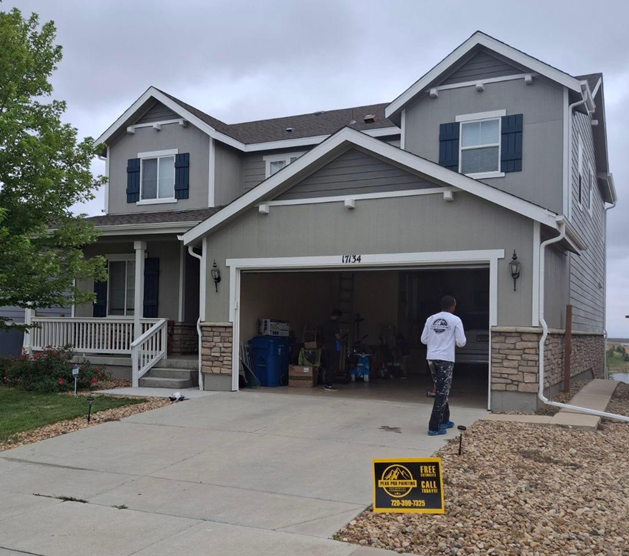 A two-story grey suburban house with a person walking into the open garage and a yard sign in the gravel front yard.