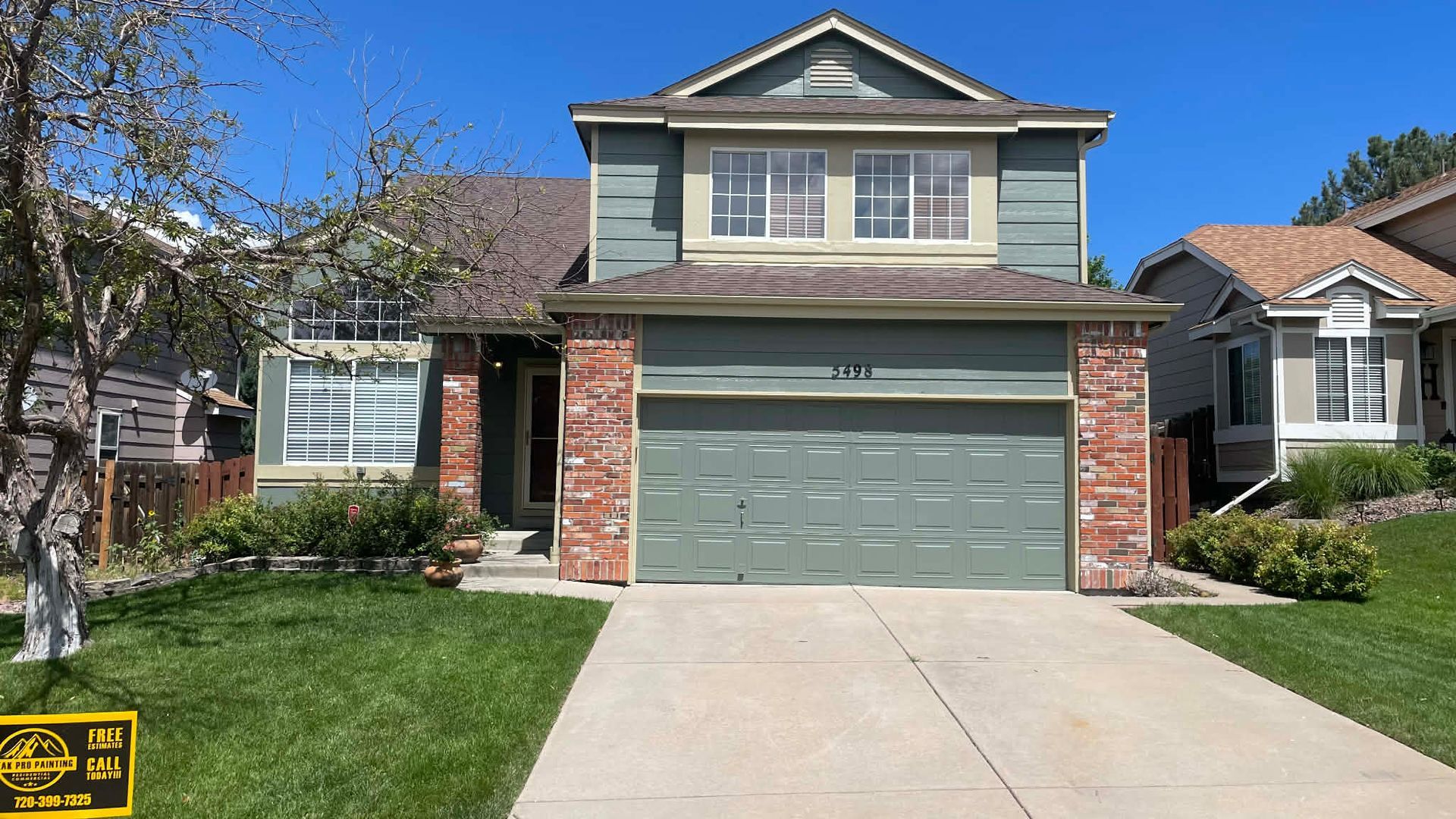 A two-story house with green siding, a brick entryway, a front-facing garage, and a green lawn under a clear blue sky.