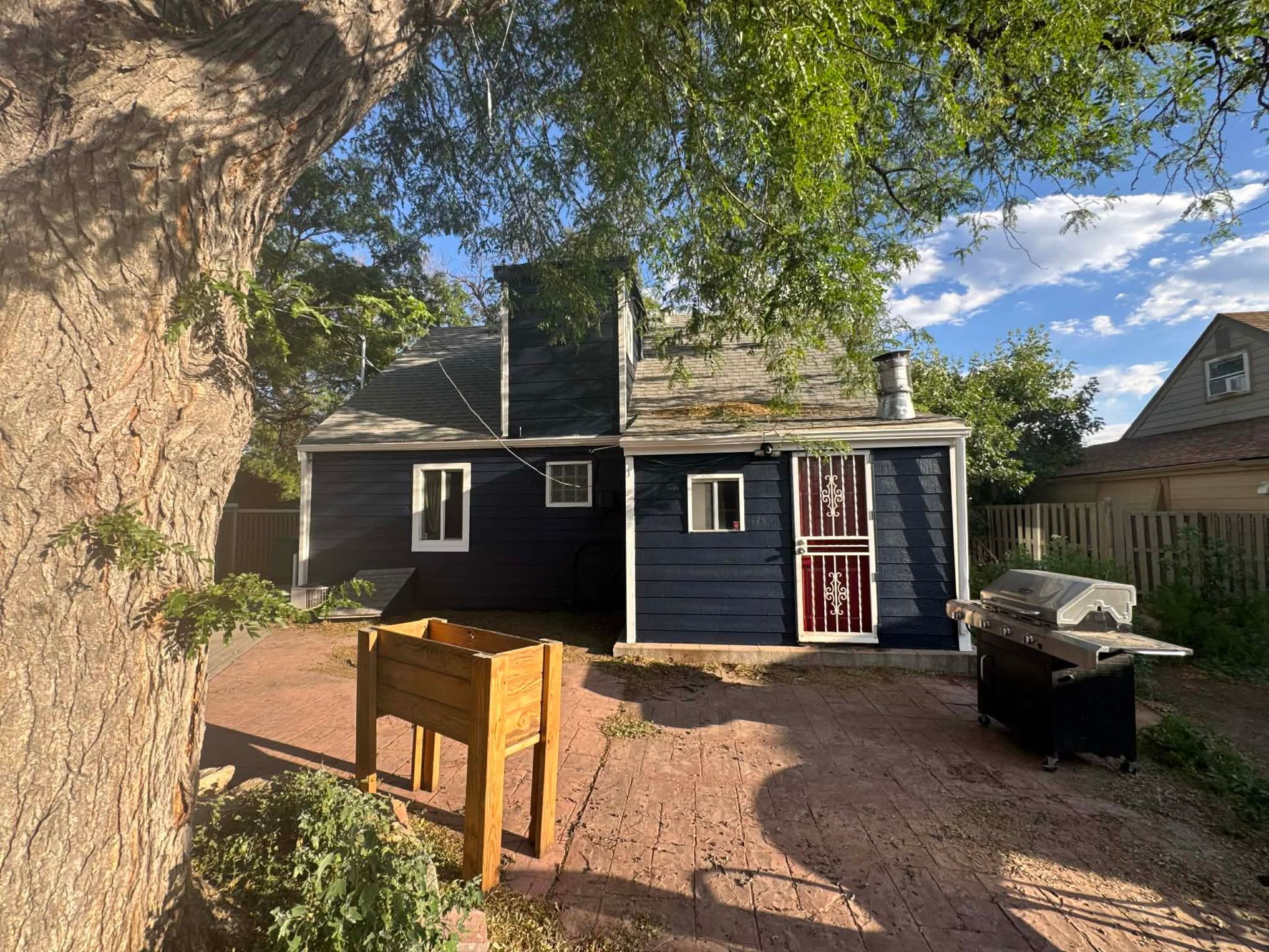 A dark blue house with white trim and a patio featuring a wooden planter and a grill, viewed from a tree-lined backyard.