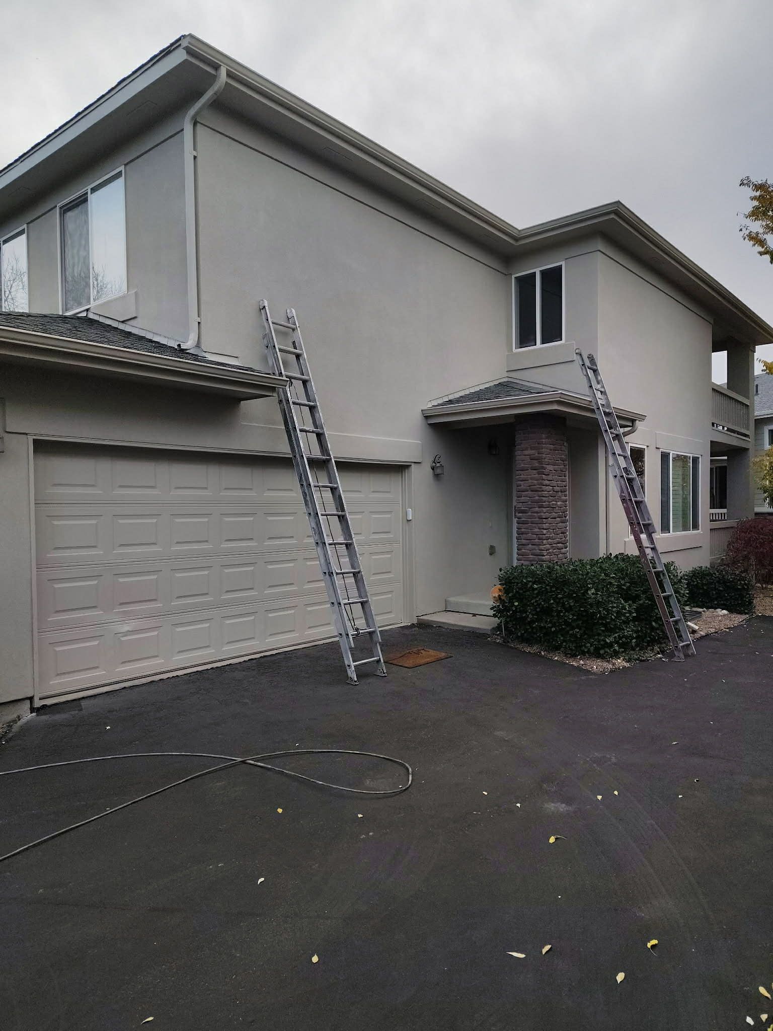 Two multi-position ladders lean against the beige stucco wall and roof of a two-story house on a cloudy day.